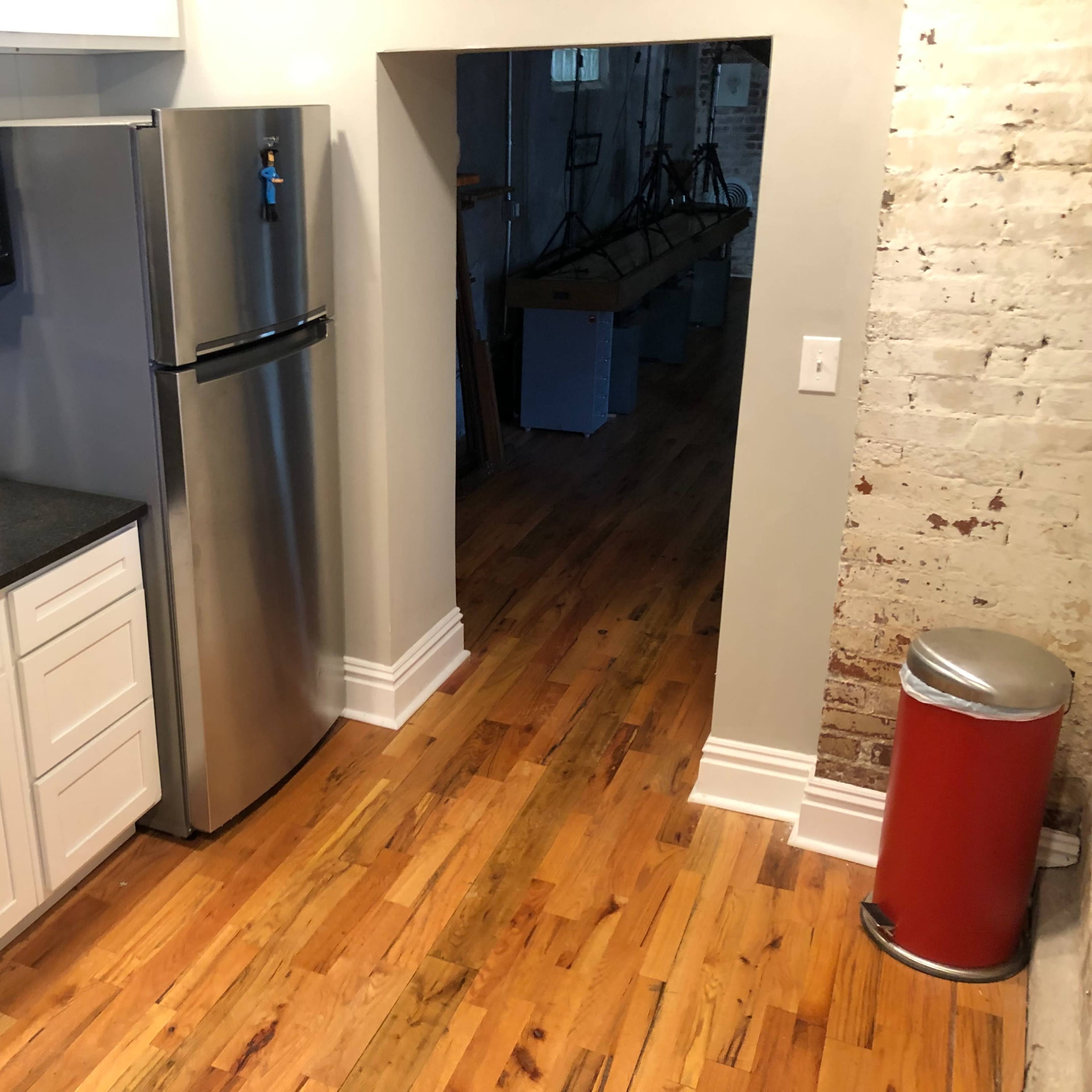 A kitchen area with stainless steel appliances, including a refrigerator and microwave, alongside white cabinets and hardwood flooring, leading to an open doorway into another room.