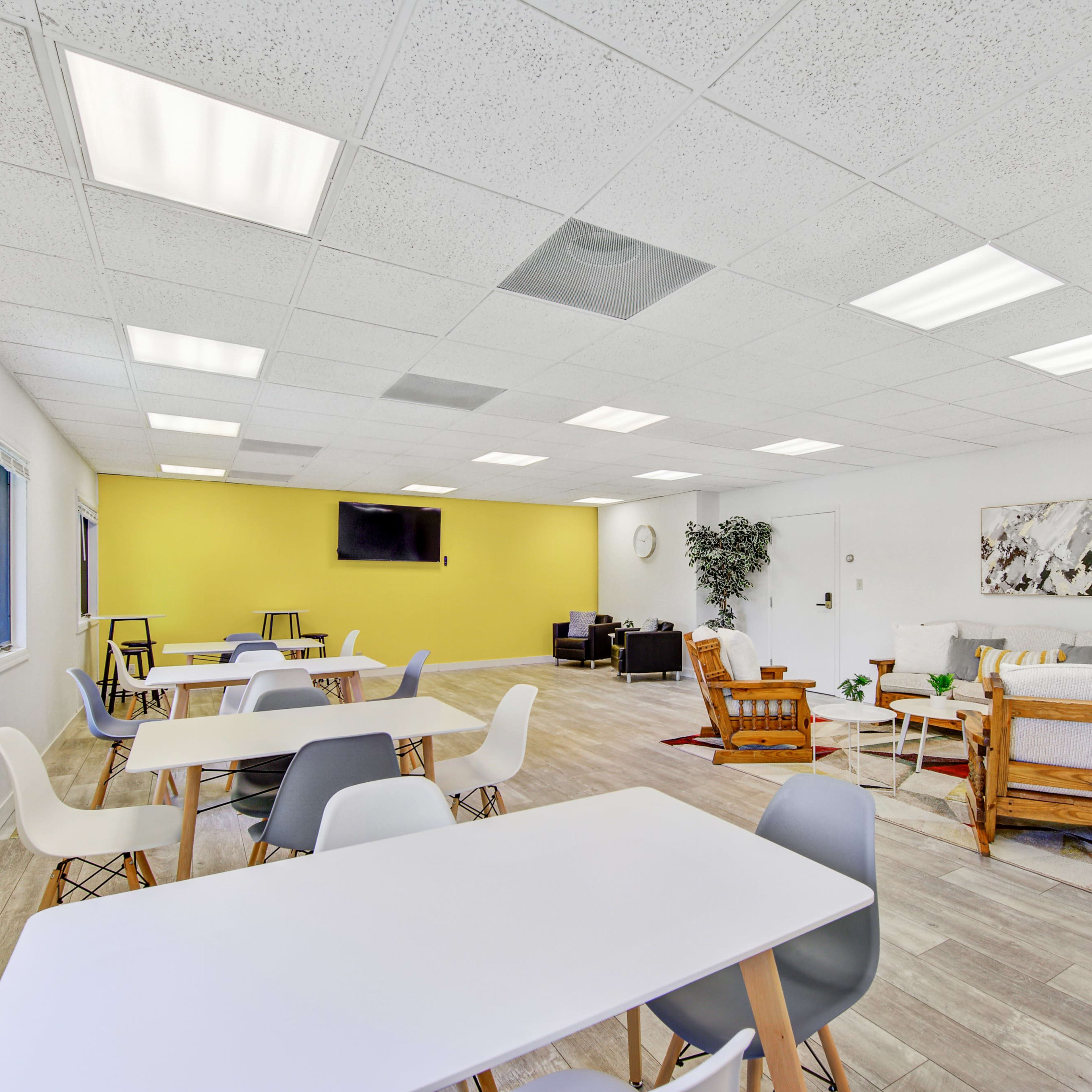 A spacious room features multiple tables and chairs, a mounted TV, and a seating area with a couch and plants against a yellow wall.