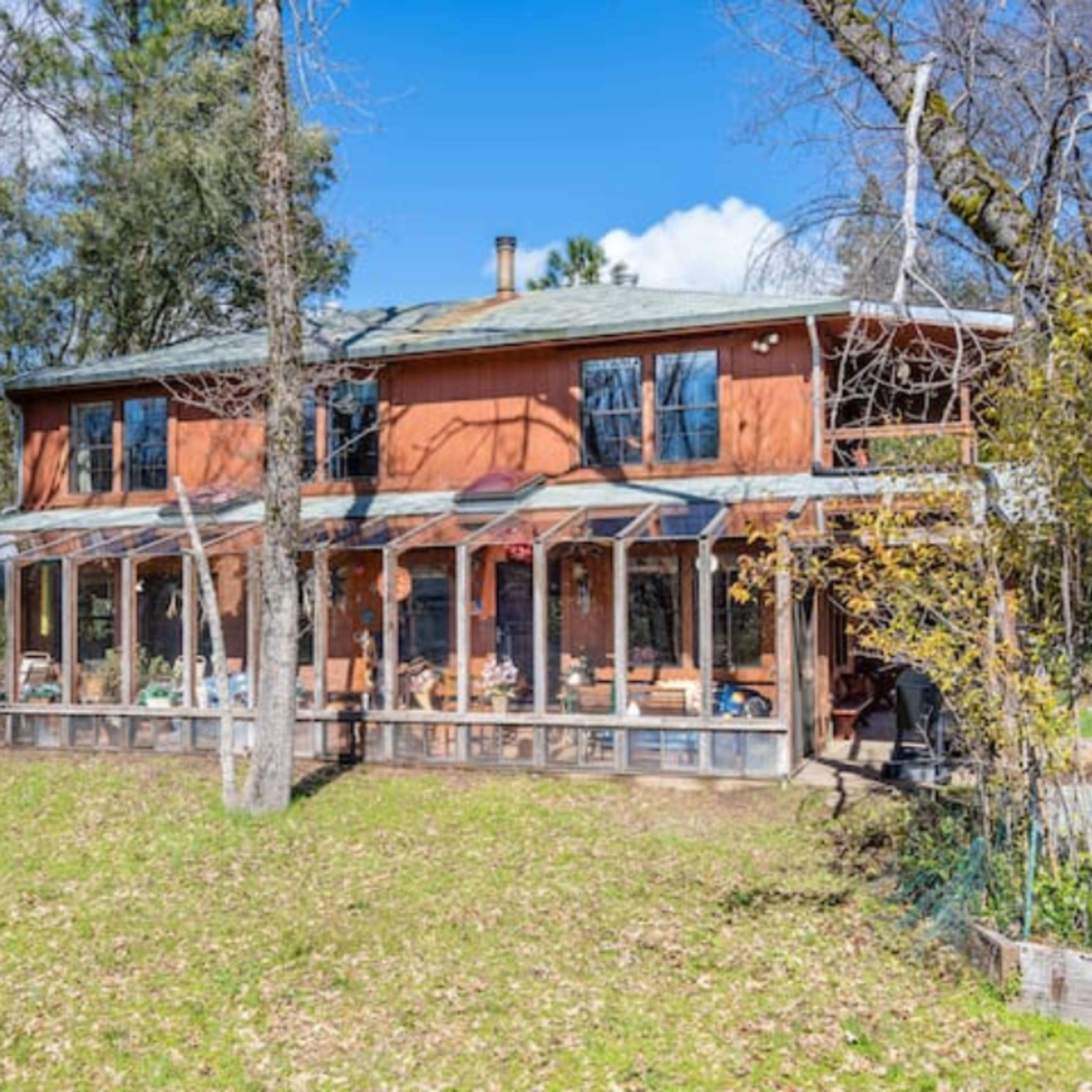 A two-story house with a reddish-orange exterior features a large glass-enclosed porch and is surrounded by trees and greenery.