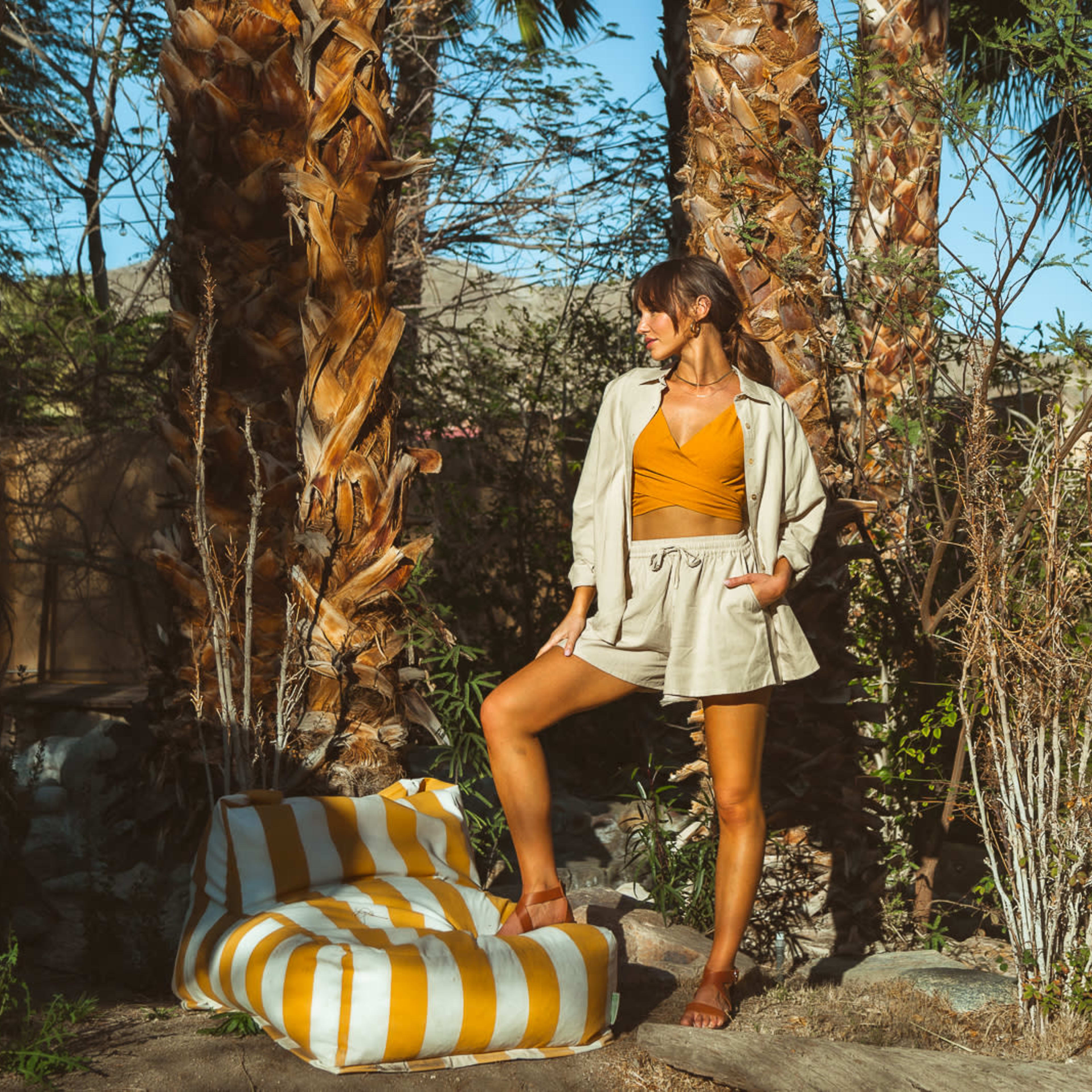 A person stands among palm trees in a garden, wearing a light-colored outfit and an orange top, with a striped outdoor cushion beside them.