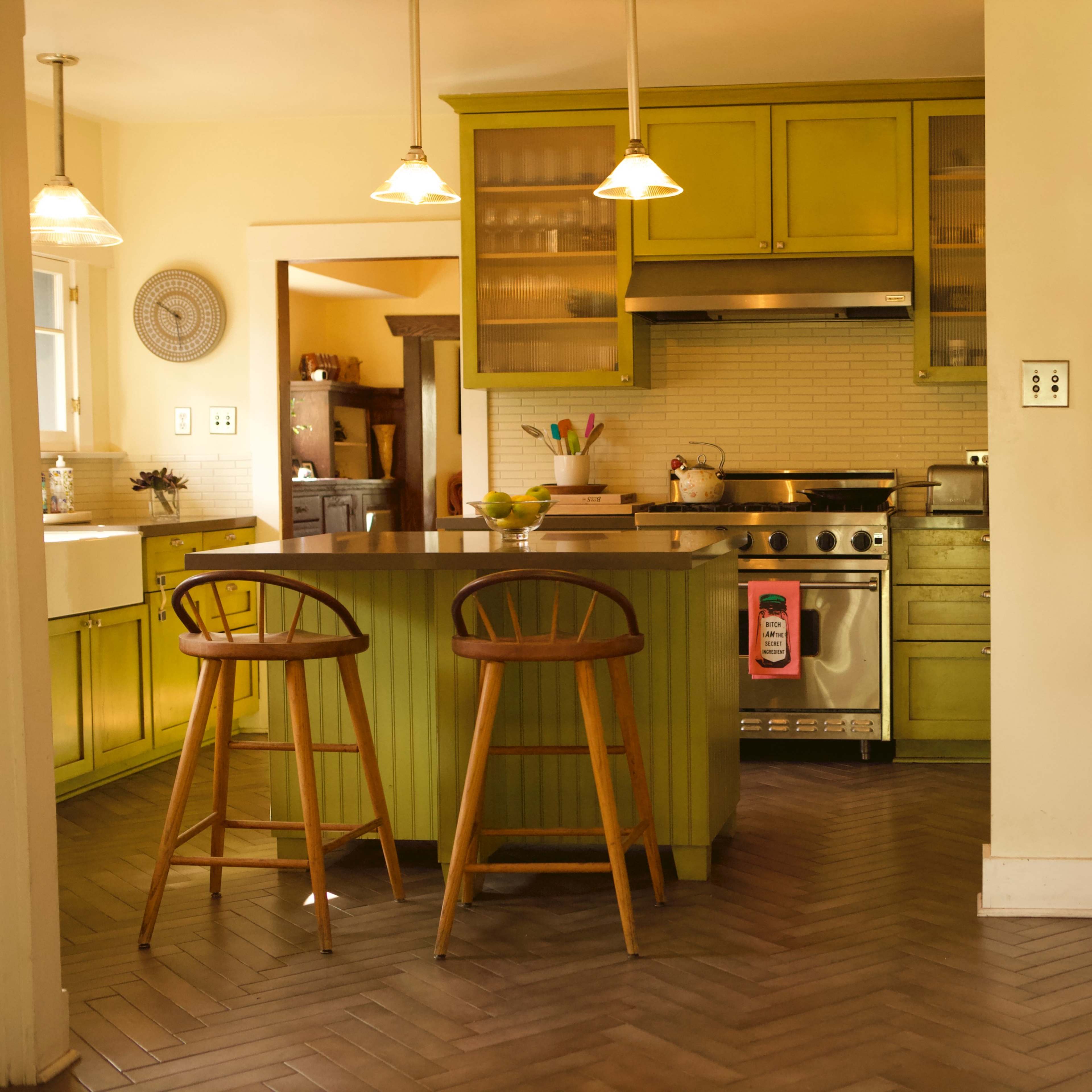 A modern kitchen with green cabinets, stainless steel appliances, and a central island with two wooden bar stools.