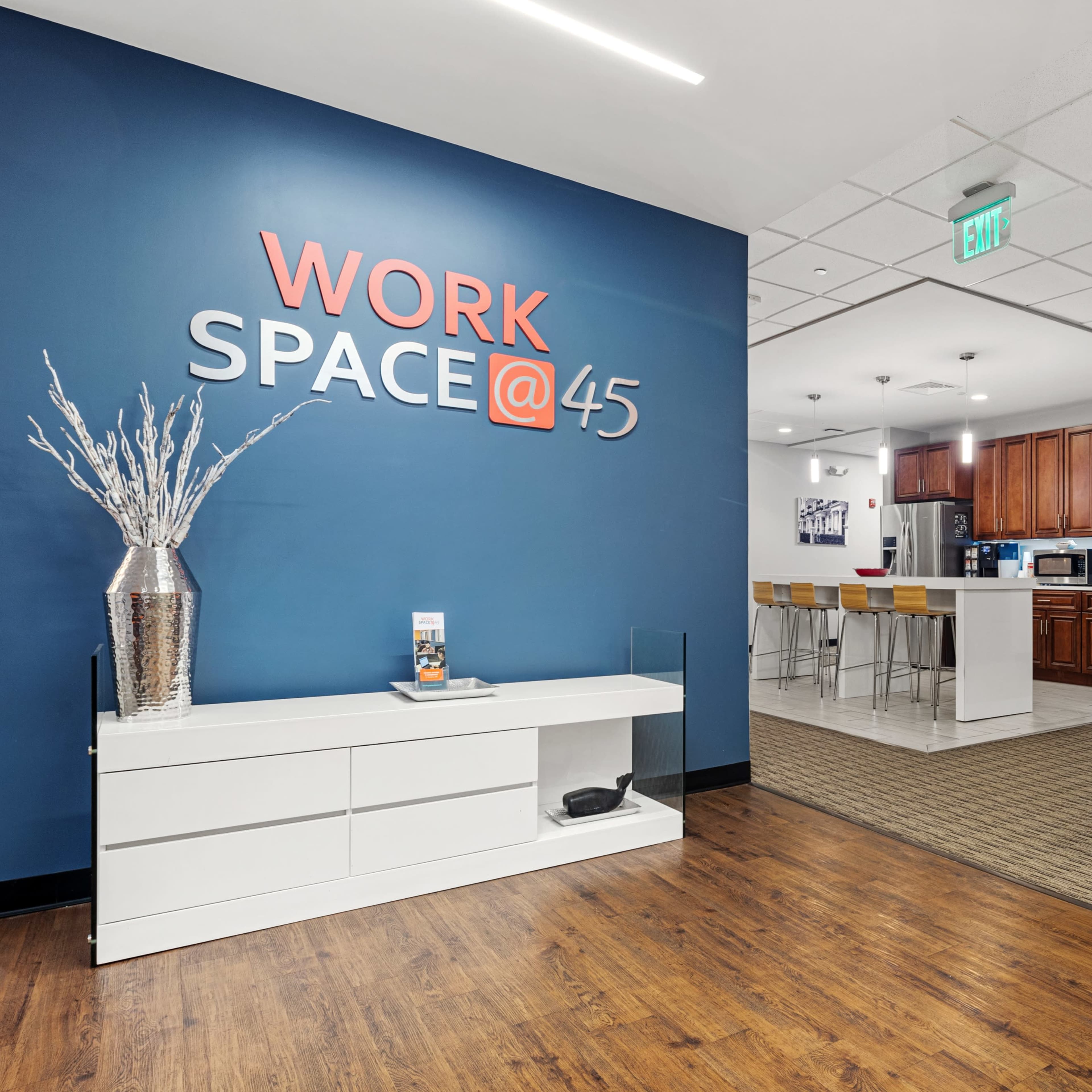 A modern office space with a blue and orange accent wall, a reception area with a sleek console table, and a kitchen area in the background.