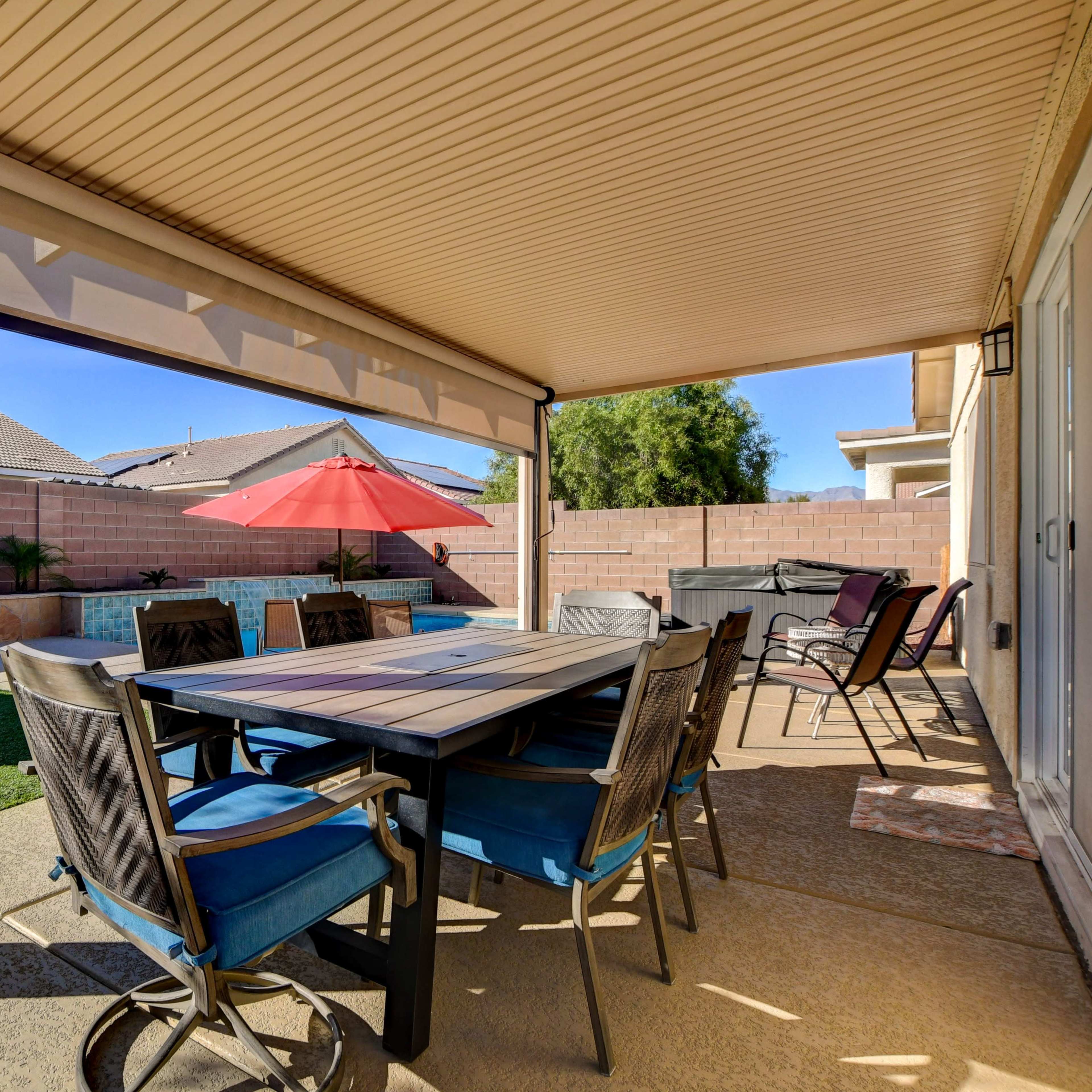 A covered patio features a dining table and chairs, overlooking a grassy area and a swimming pool with an umbrella in the background.