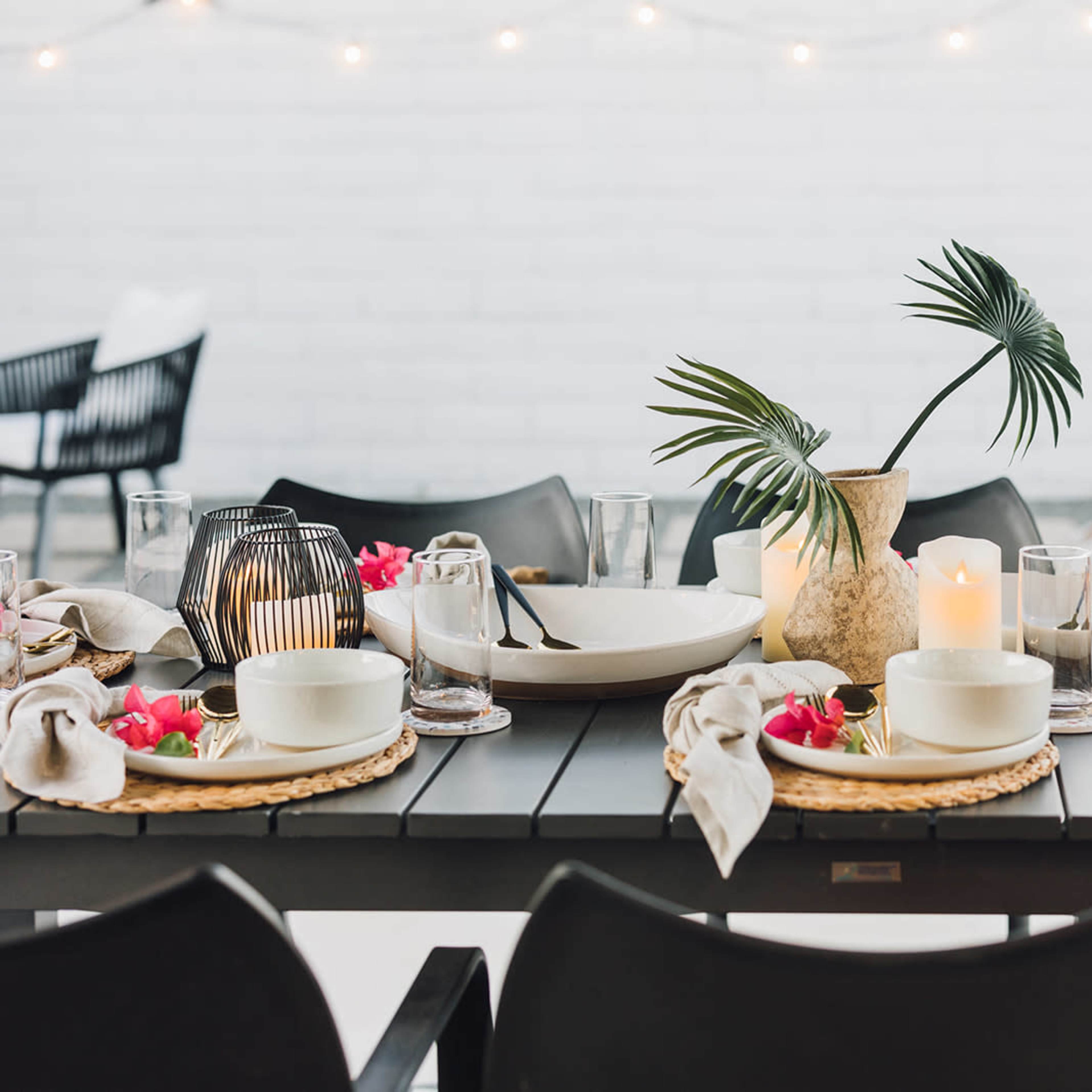 A dining table is set with plates, glasses, and decorative candles, accompanied by a vase with greenery and flowers.