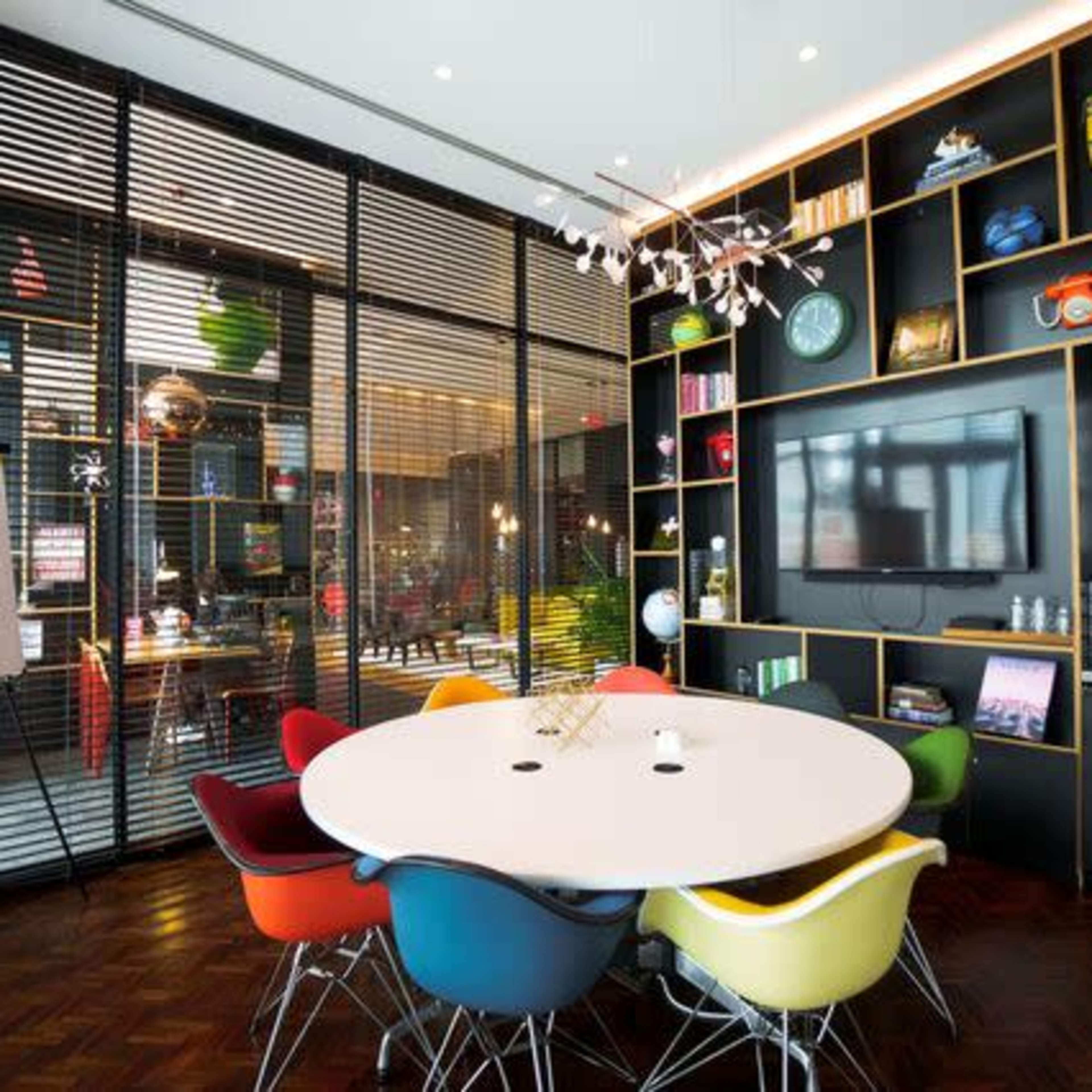A modern conference room features a round table surrounded by colorful chairs, with a wall-mounted television and shelves displaying decorative items.