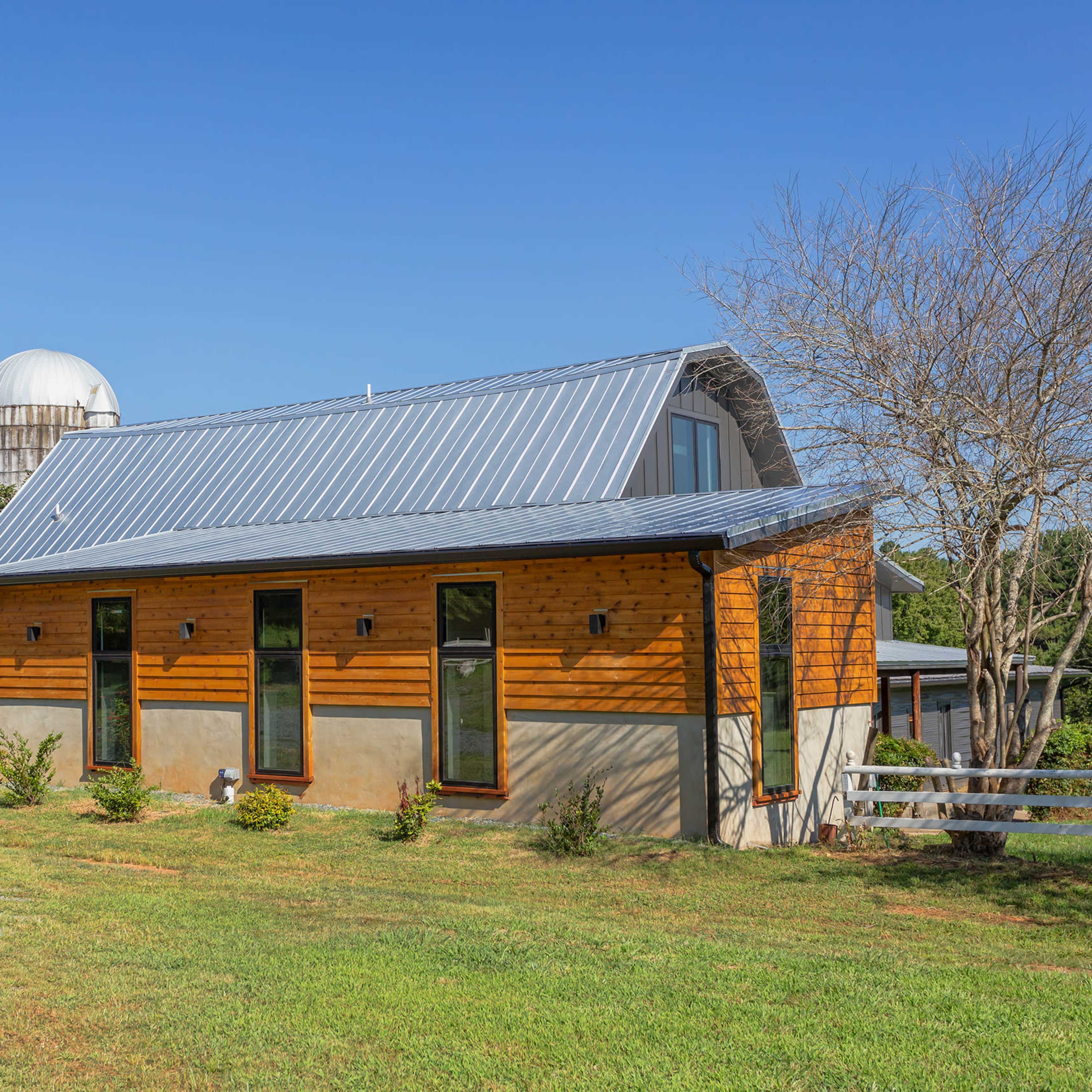 A modern farmhouse with a metal roof and wooden accents is set against a clear blue sky, surrounded by green grass and a silos in the background.