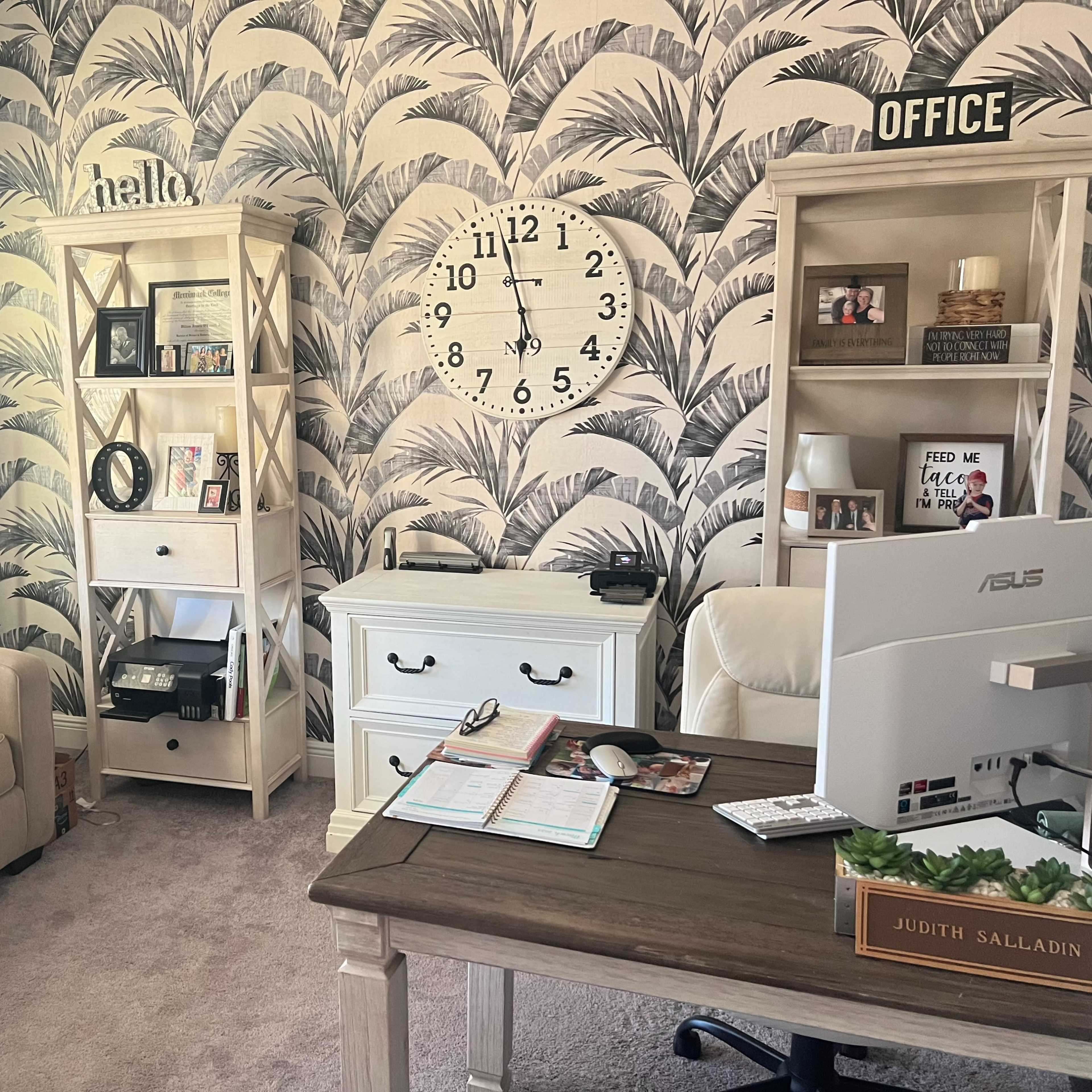 A home office featuring a desk with a computer, bookshelves, and a decorative wall with a tropical leaf pattern.