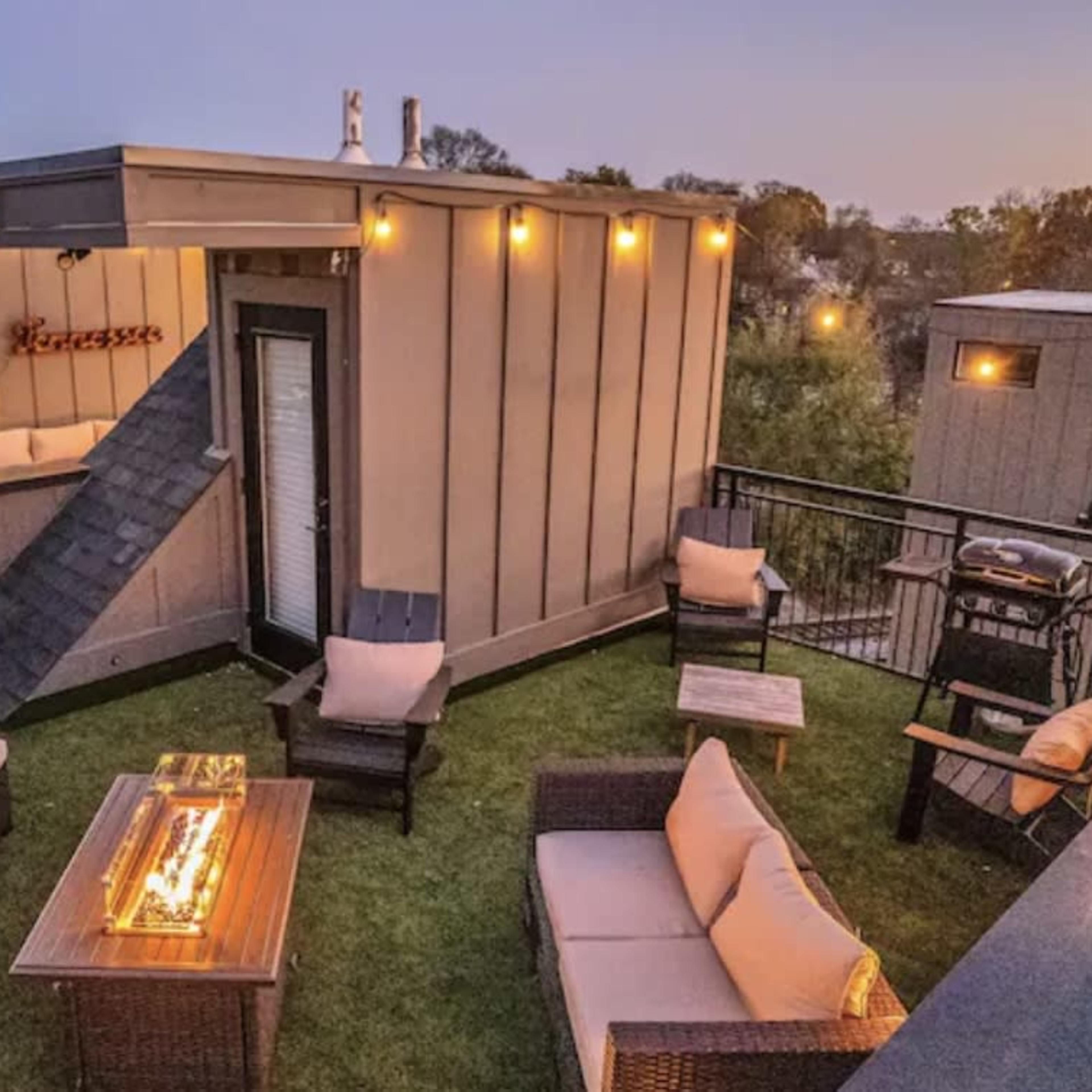 A rooftop lounge area with cozy seating arrangements, a grill, and decorative lighting at dusk.
