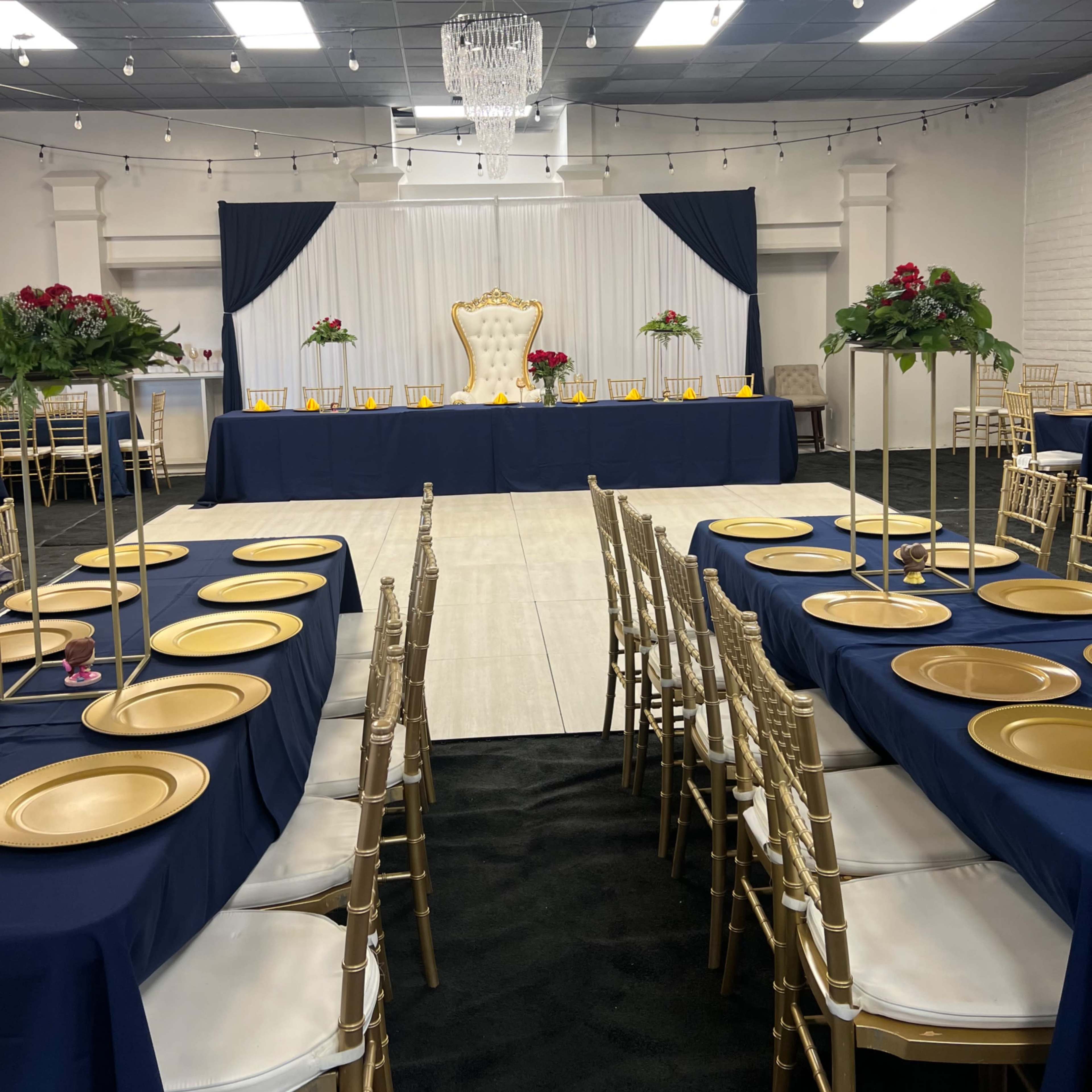 A banquet hall set up with gold and navy table settings, featuring a raised backdrop with a throne and floral arrangements at the front.