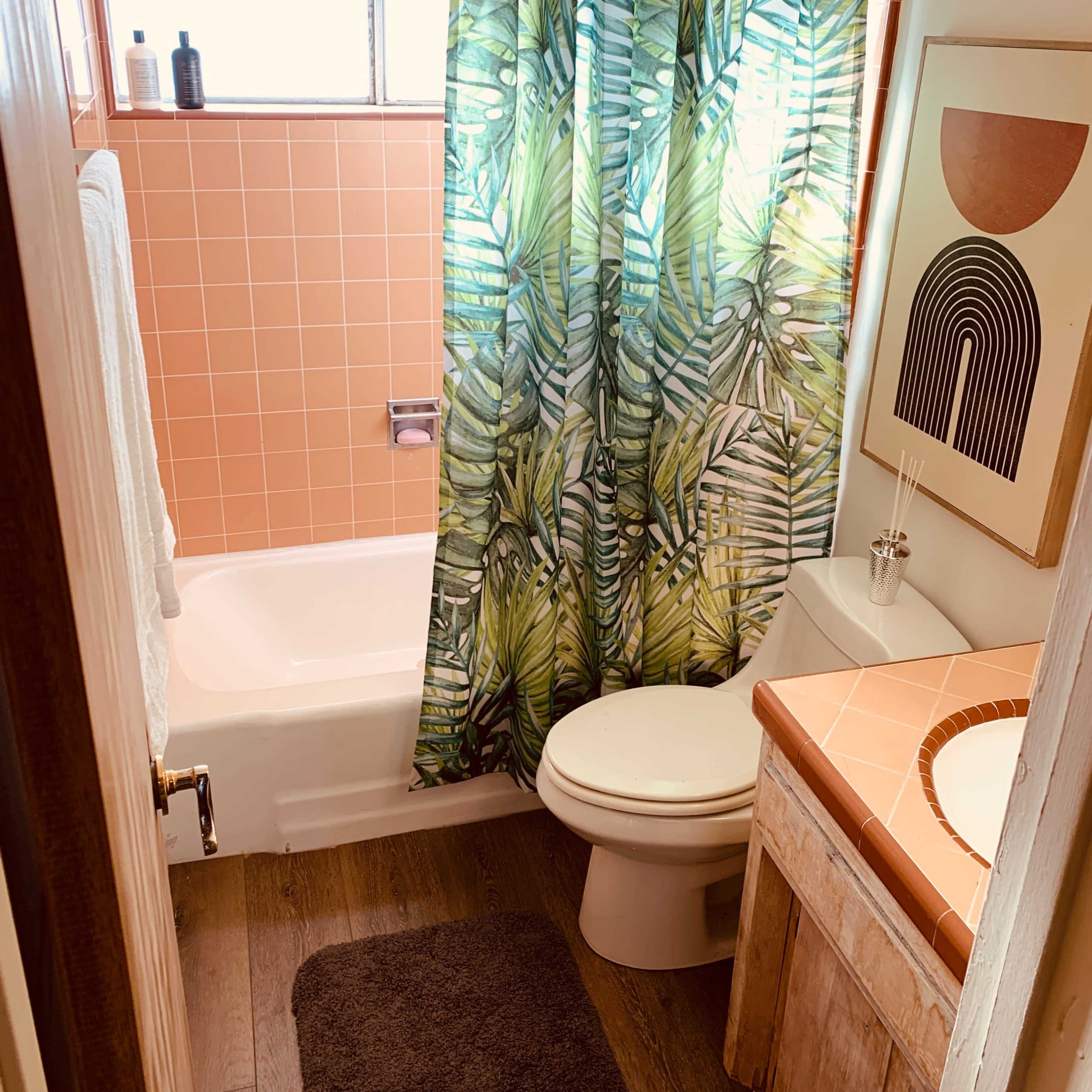 The bathroom features a bathtub with a leafy shower curtain, a toilet, a sink with a tiled countertop, and a window allowing natural light.