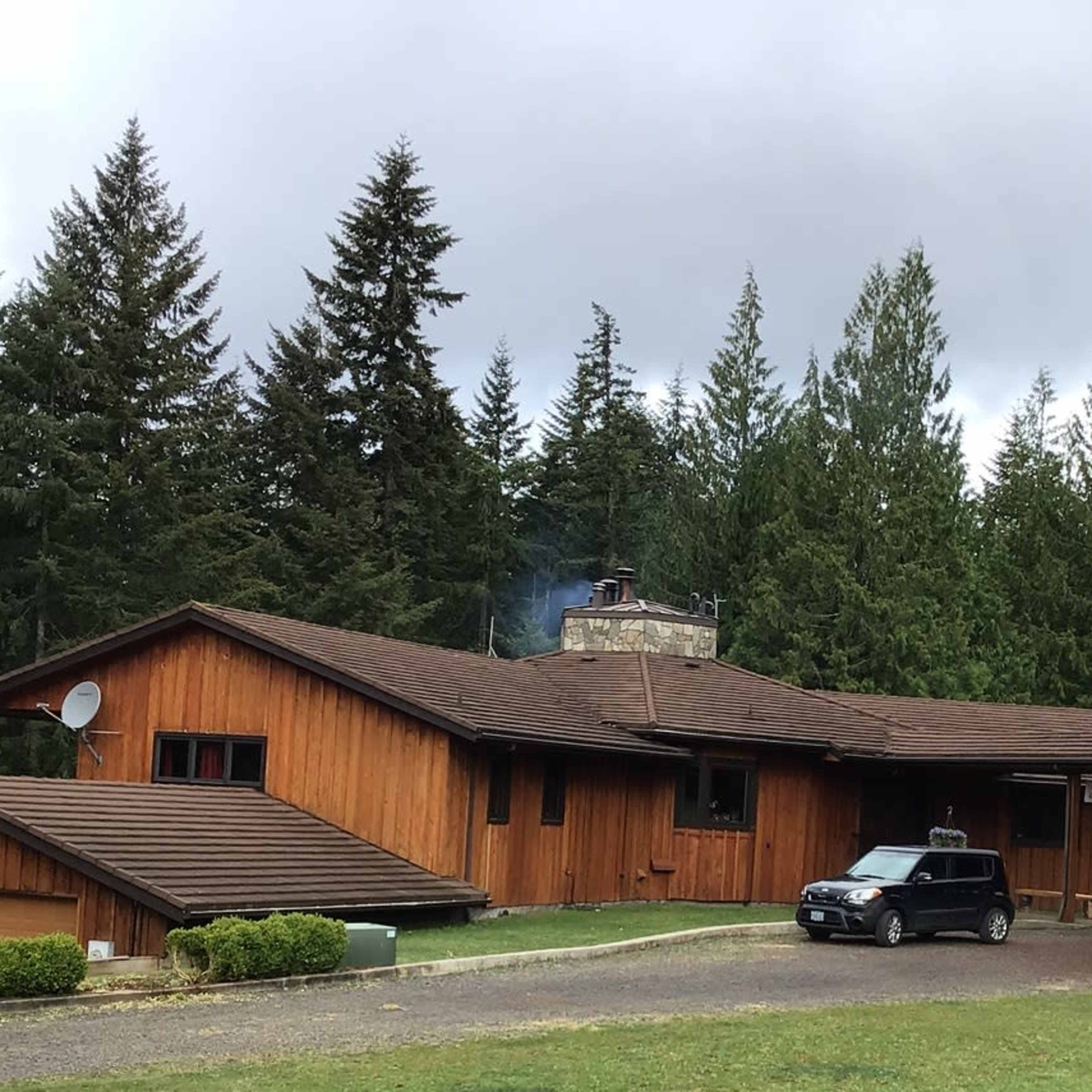 A wooden house with a stone chimney sits amidst tall trees, with a small car parked in front on a gravel drive.