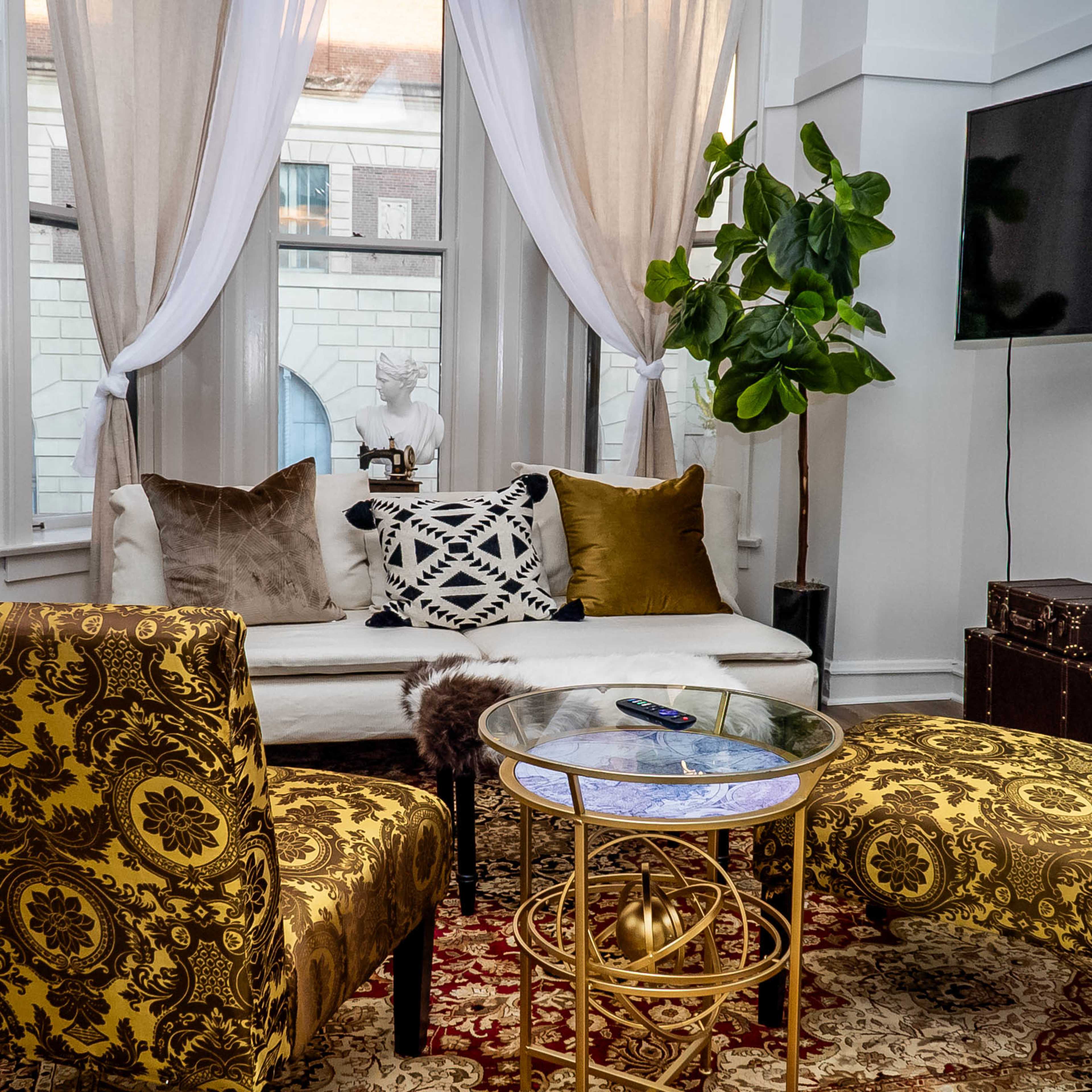 A cozy living room with a white sofa, two ornate yellow chairs, a glass coffee table, and a potted plant in the corner.