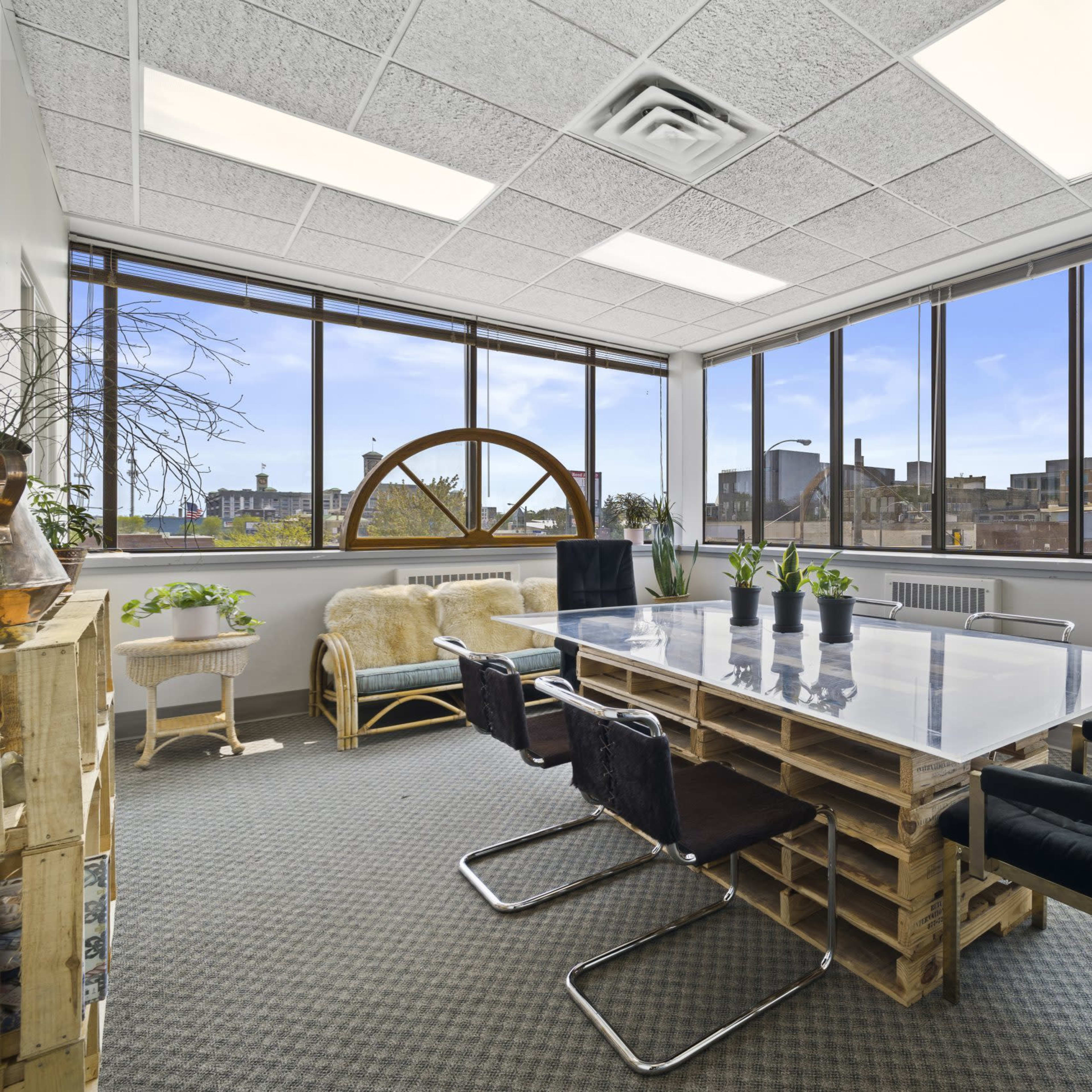 A bright office meeting room with large windows, featuring a glass table surrounded by black chairs and various plants.