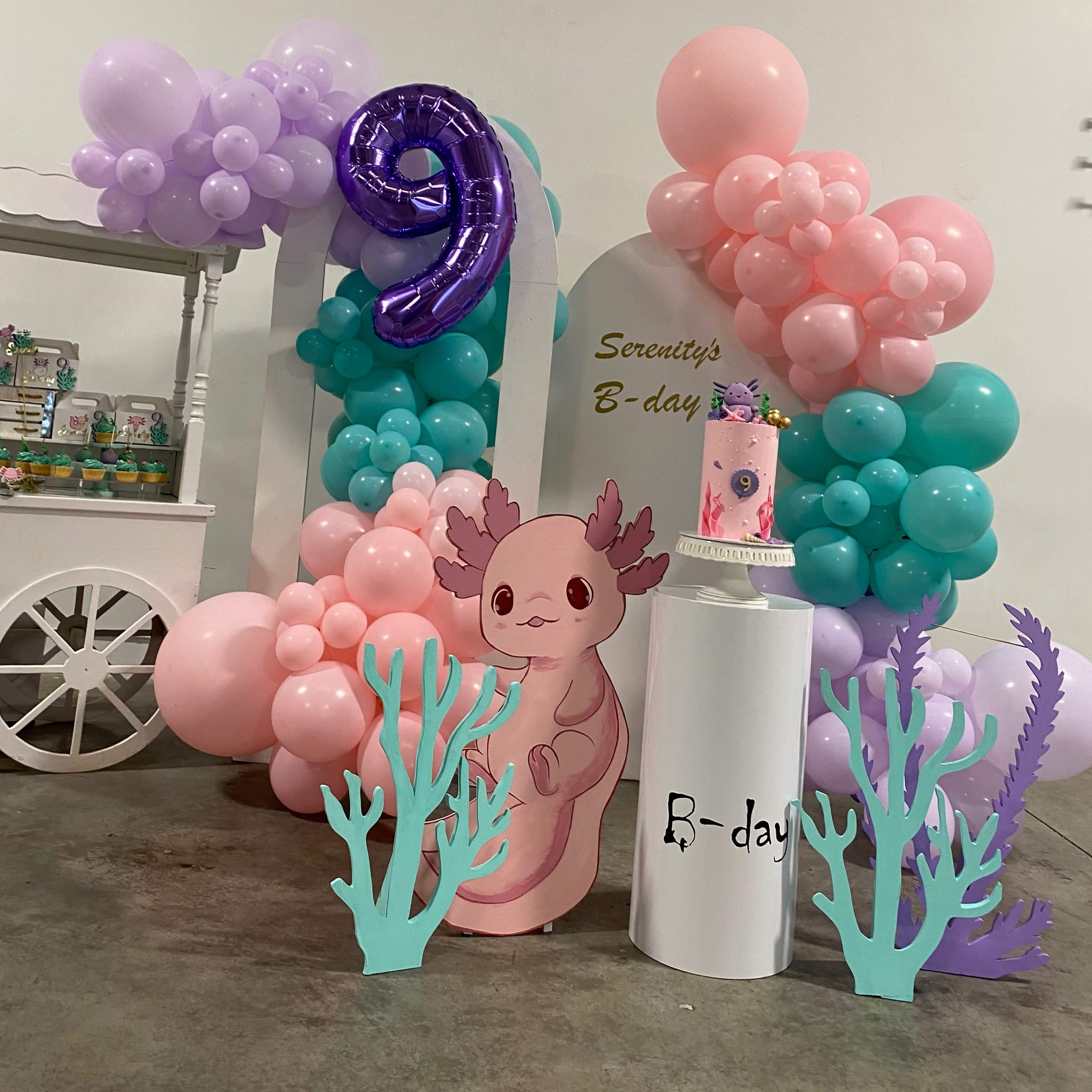 A colorful birthday decoration setup with balloons in shades of pink, purple, and blue, a birthday cake, and a cartoon axolotl figure.