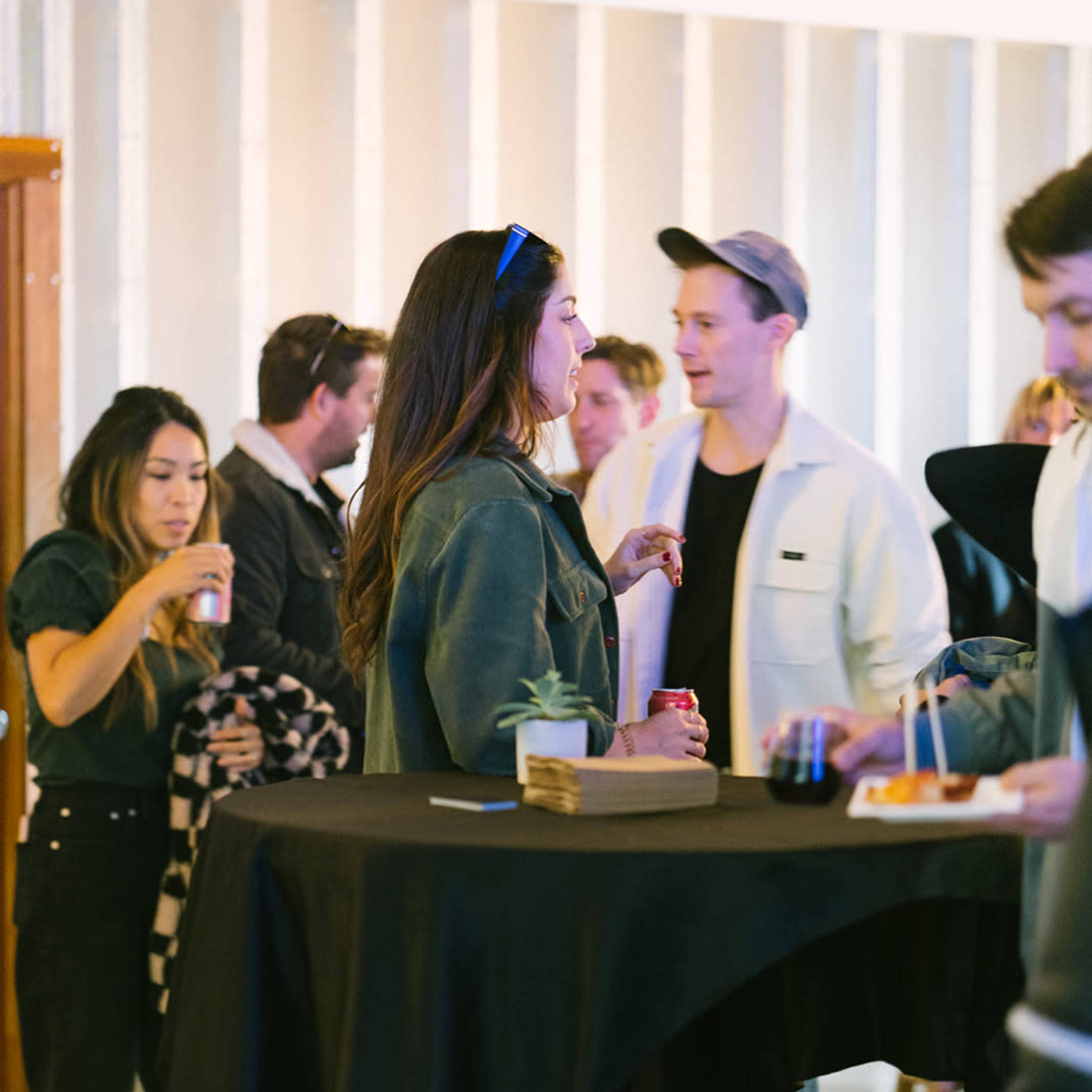 A group of people socializes around high tables at an event, with some holding drinks and snacks.