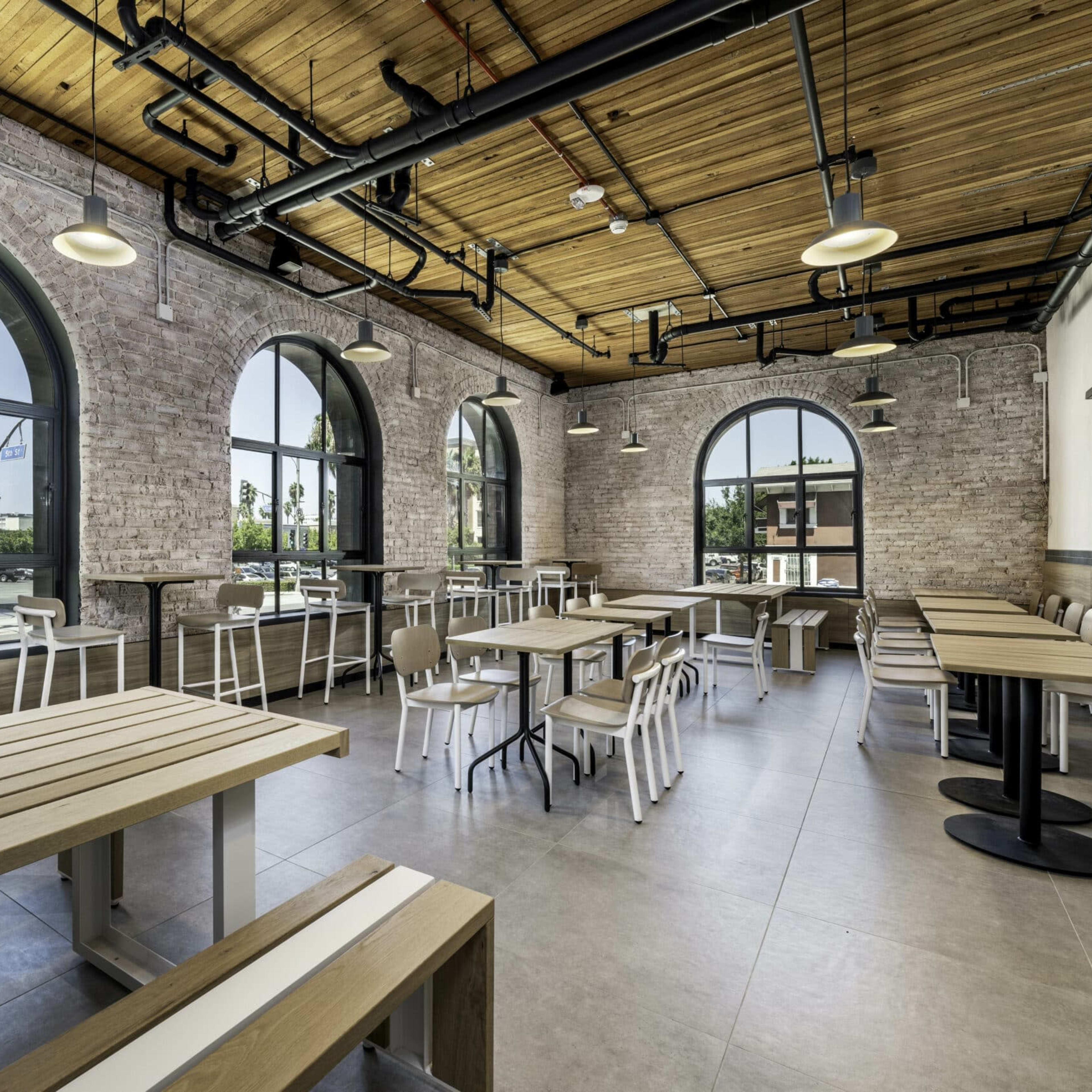A spacious, modern café interior with large arched windows, wooden ceilings, and a mix of tables and chairs arranged neatly.