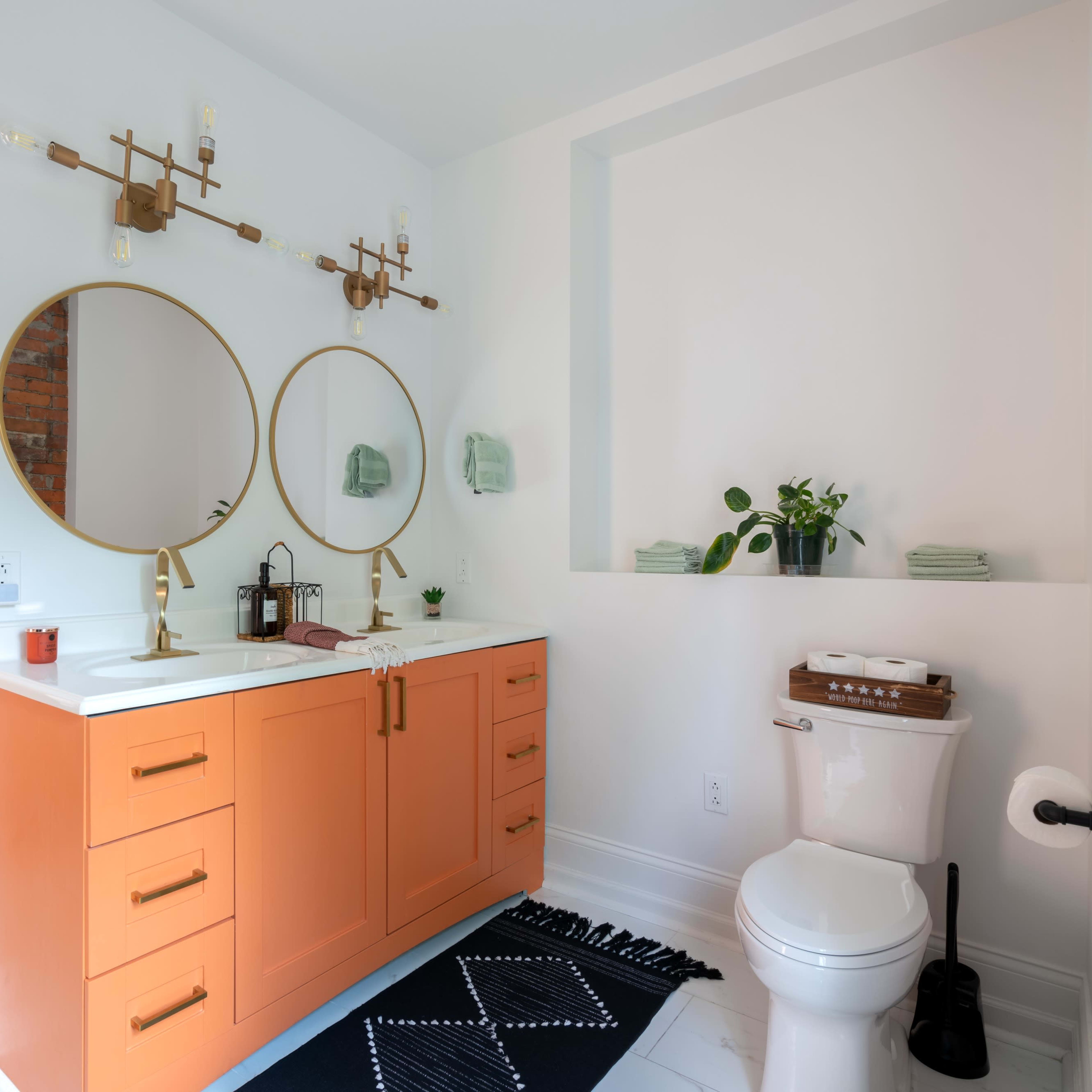 A modern bathroom featuring two round mirrors above a double sink with orange cabinetry, a toilet, and decorative plants against a white and brick wall.