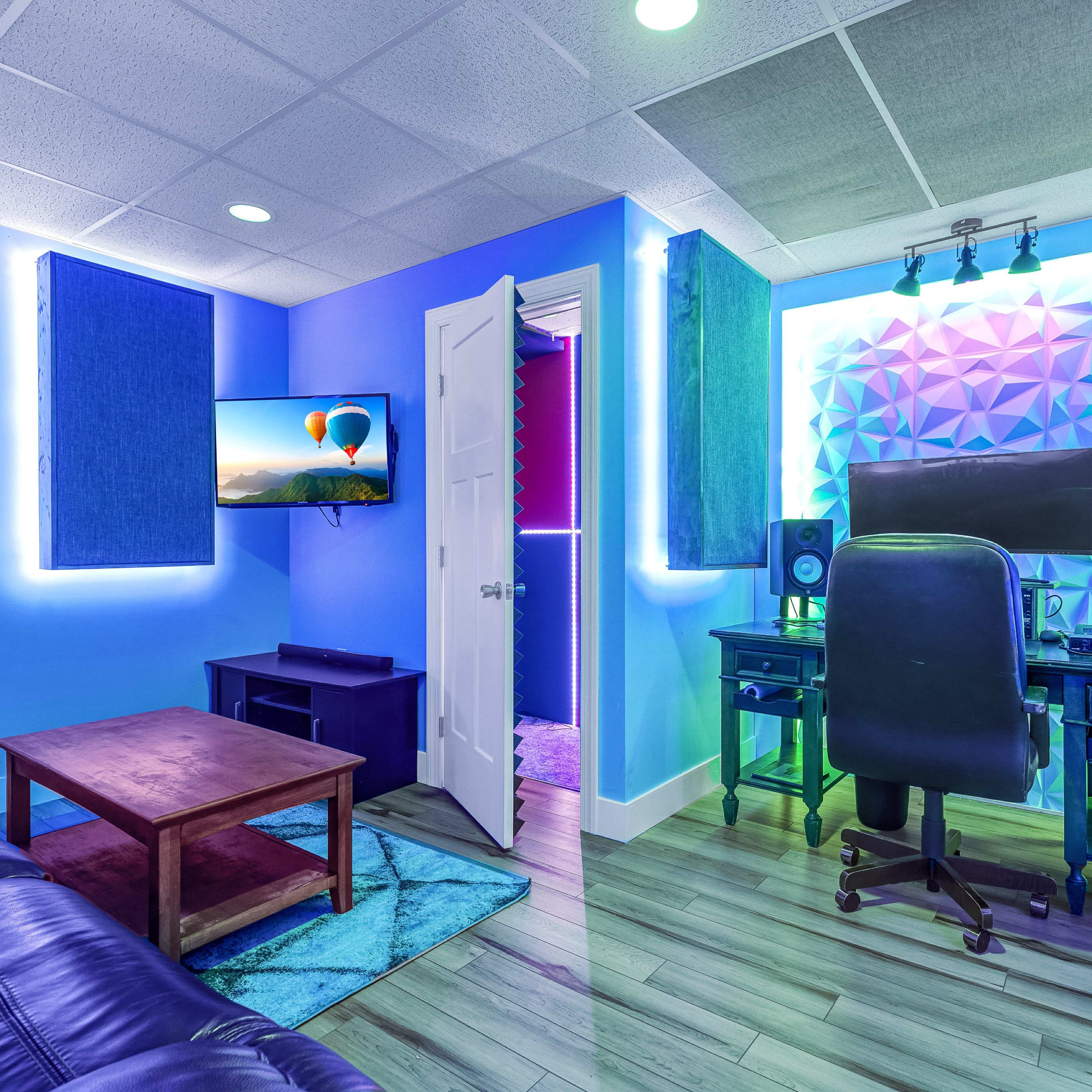 A modern room with a gaming setup, featuring a desk with monitors, a black ergonomic chair, blue walls, and acoustic panels, along with a couch and a television.