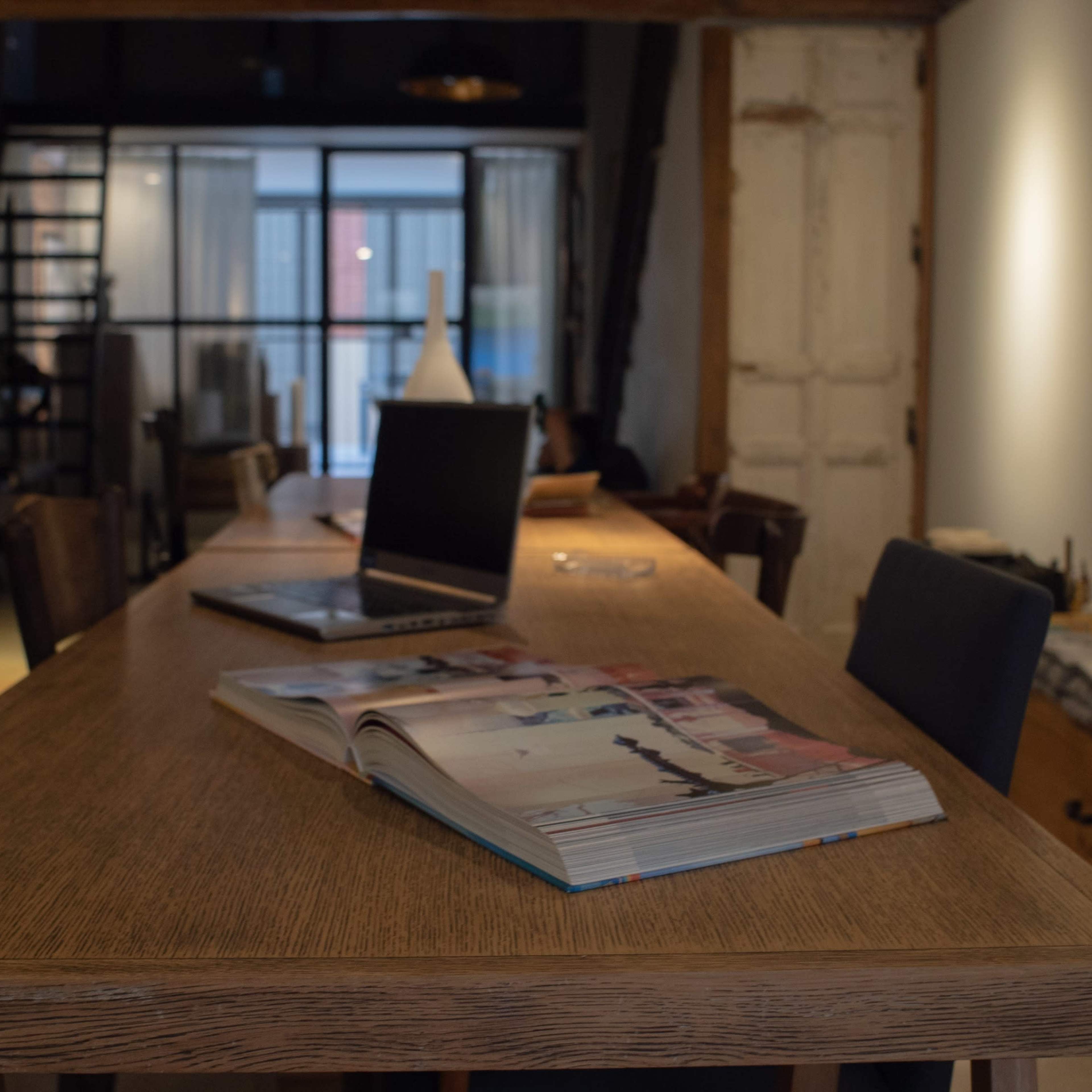 A long wooden table sits in a dimly lit room, with a laptop and an open magazine on its surface.