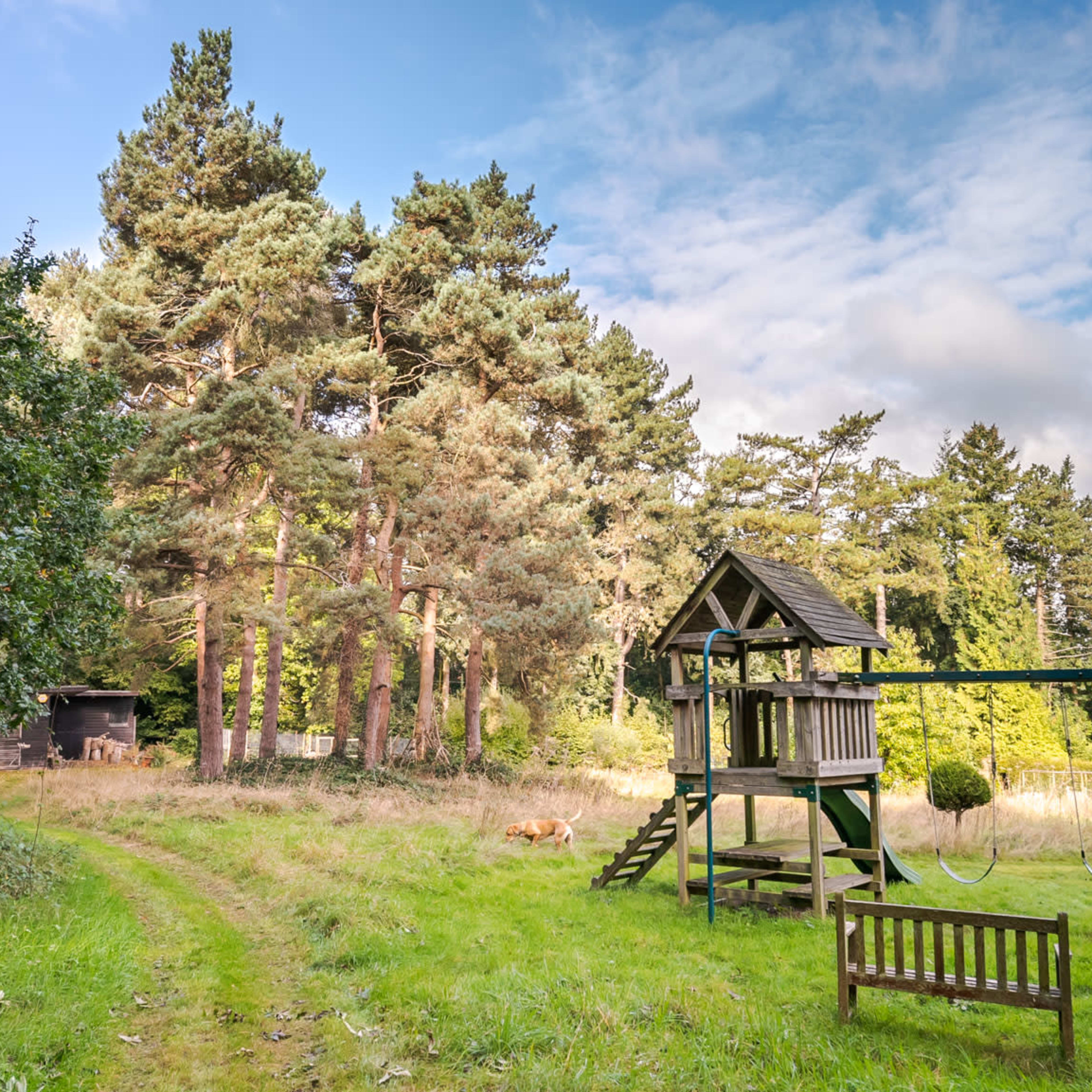 A wooden playhouse with swings is positioned near a wooded area with tall trees and a grassy pathway.