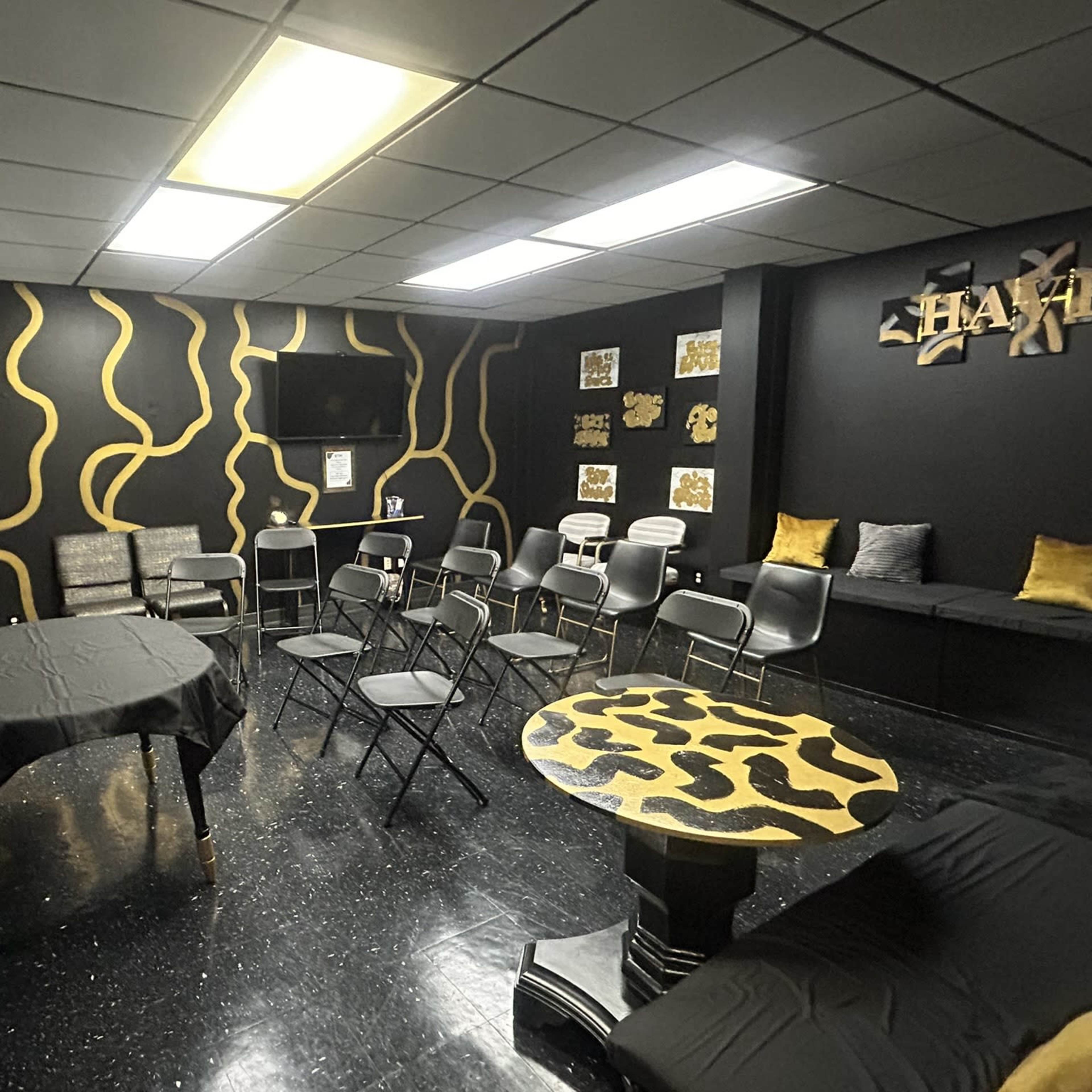 The room features black walls with gold abstract designs, a central table with black cloth, folding chairs arranged around it, and a television on one wall.