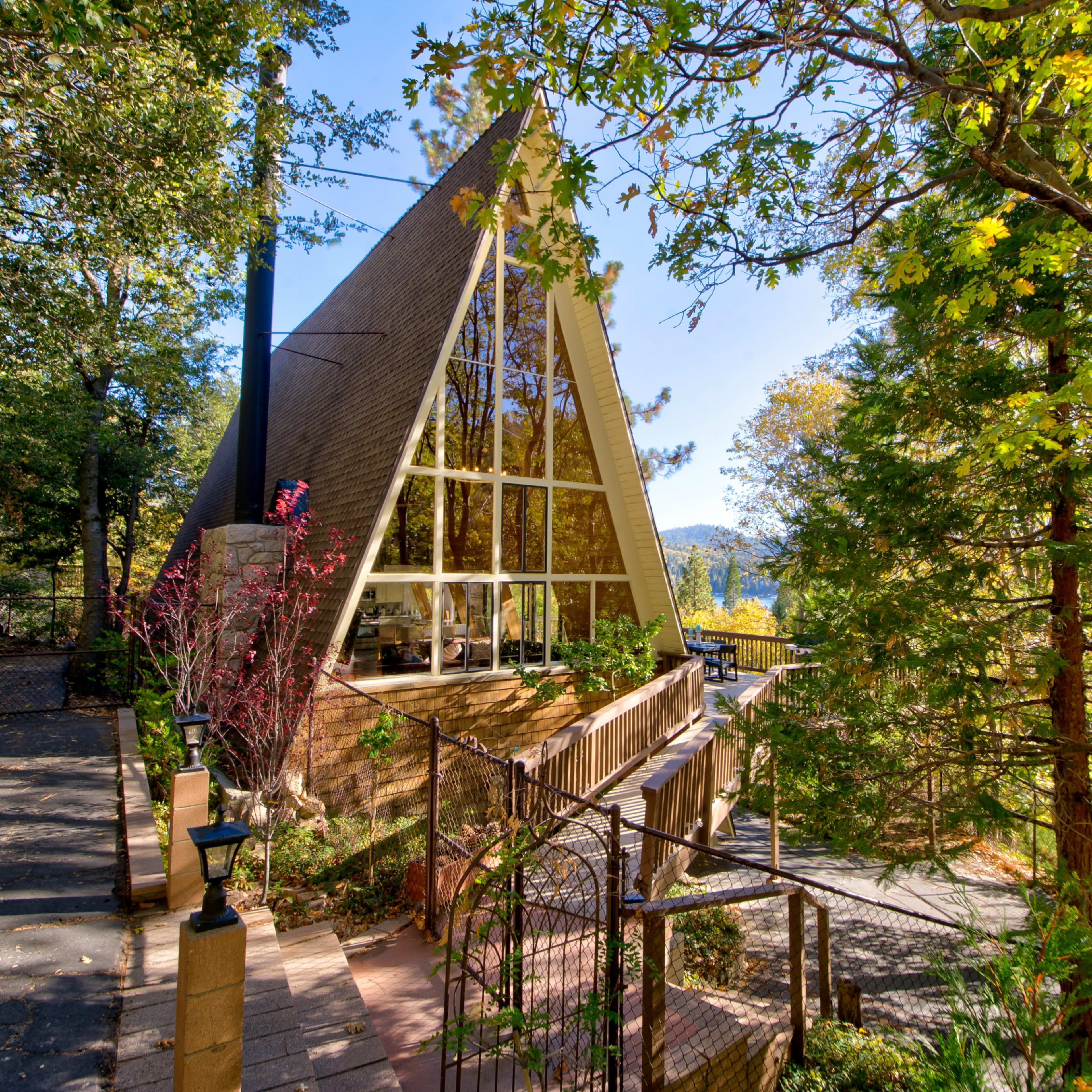 A modern A-frame house with large windows, surrounded by trees and a pathway leading to its entrance.