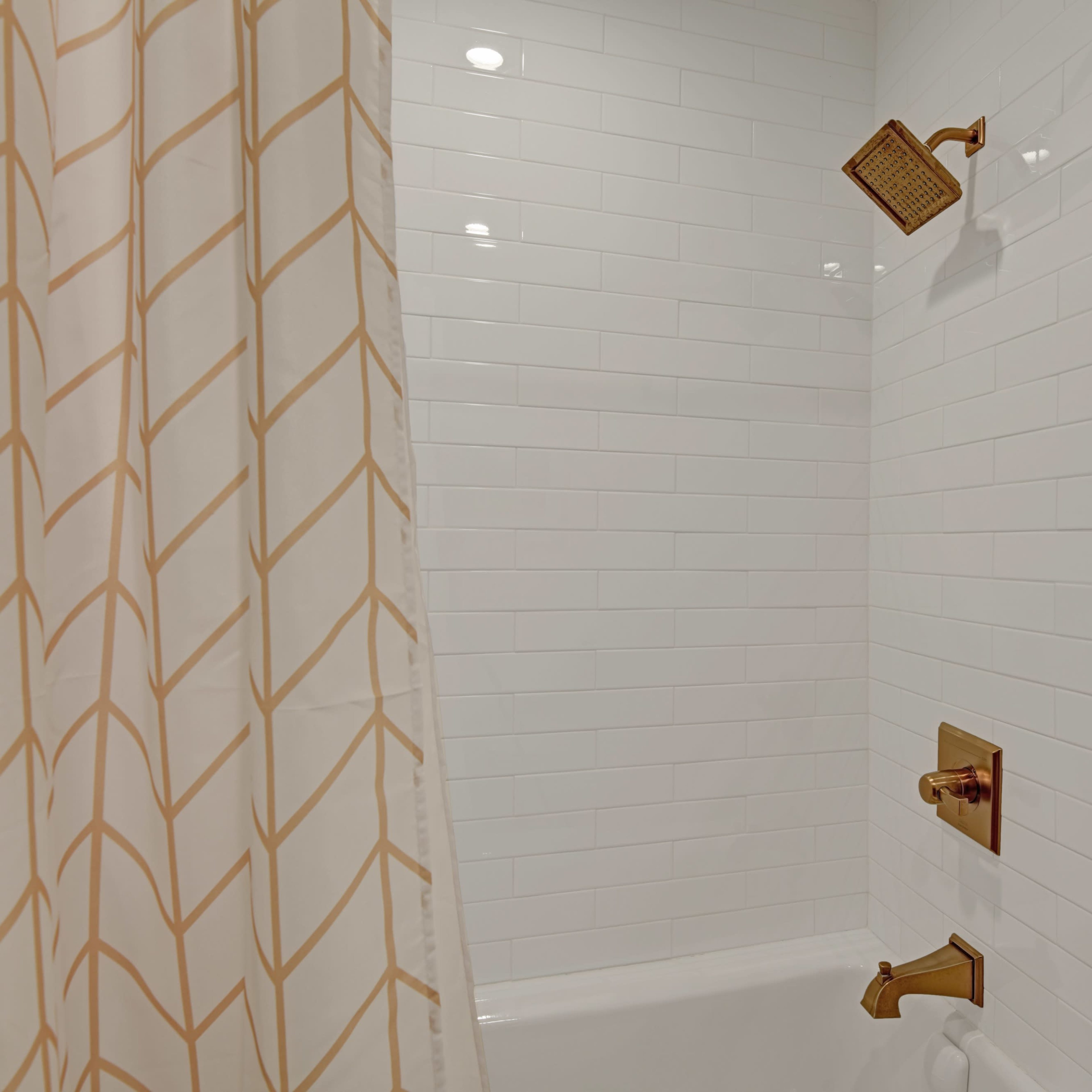 A shower area with white tiled walls, a gold showerhead, and a beige patterned shower curtain.