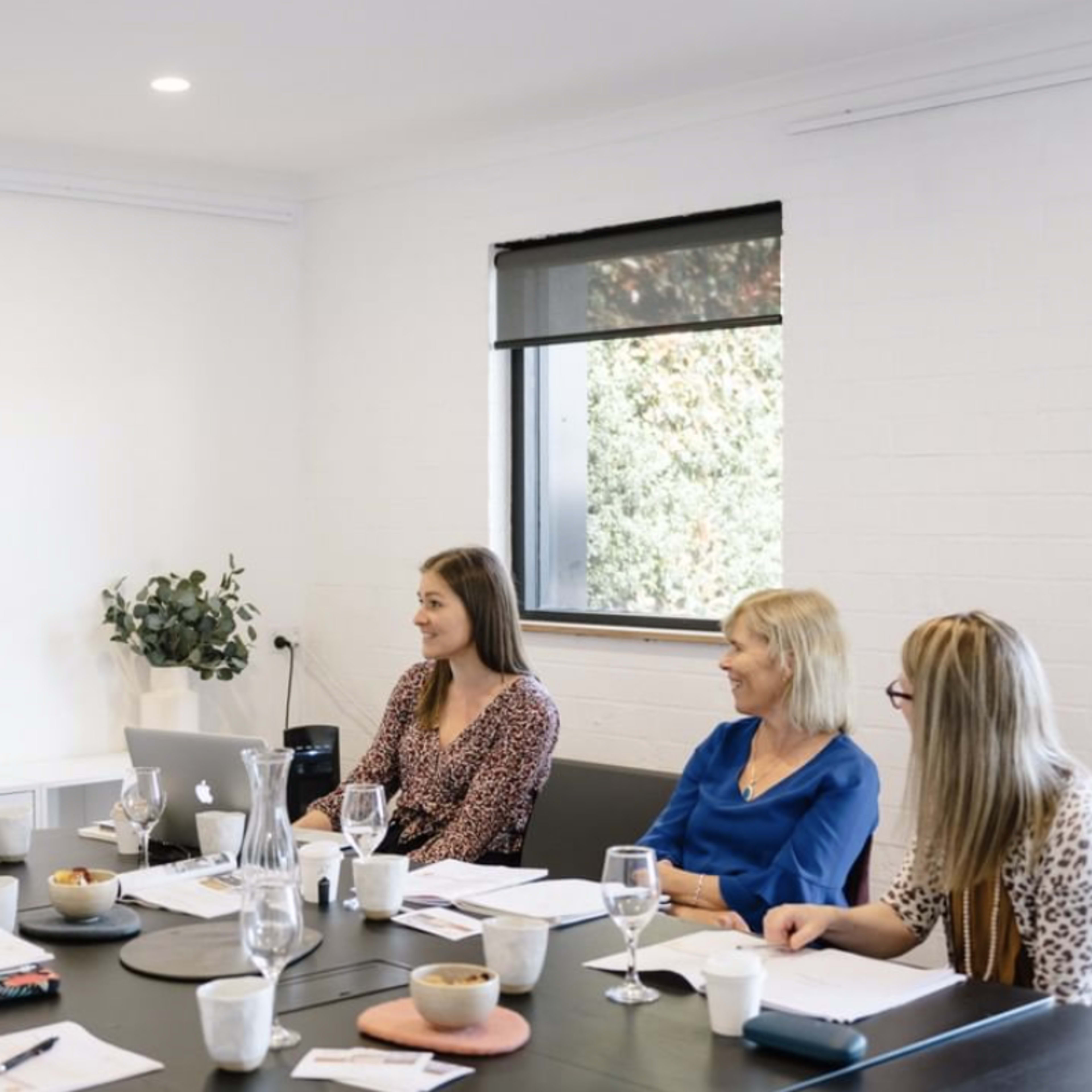 A group of five women are seated around a conference table, engaged in a discussion with laptops and documents in front of them.