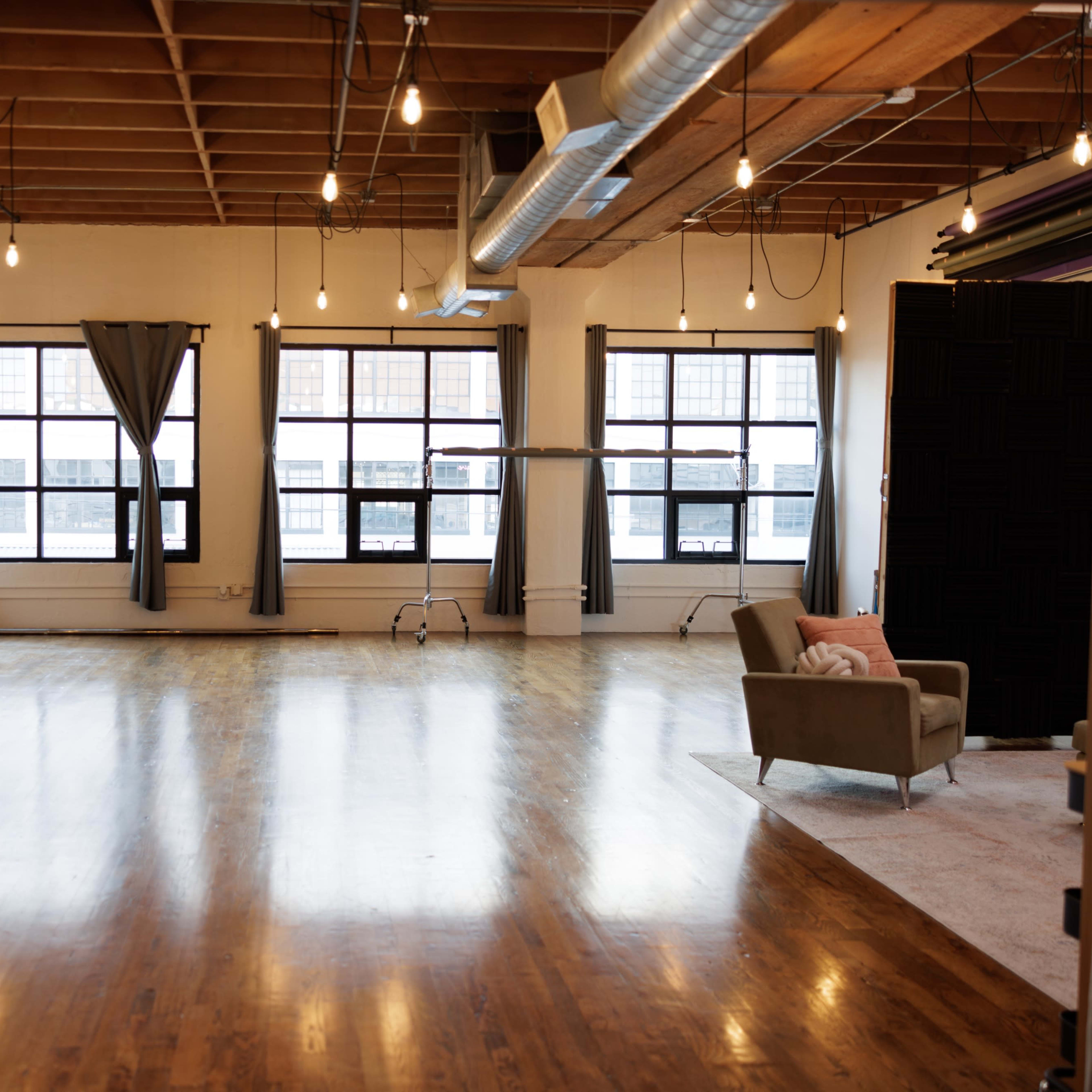 A spacious, well-lit studio features large windows, polished wooden floors, a sofa, and simple decor.