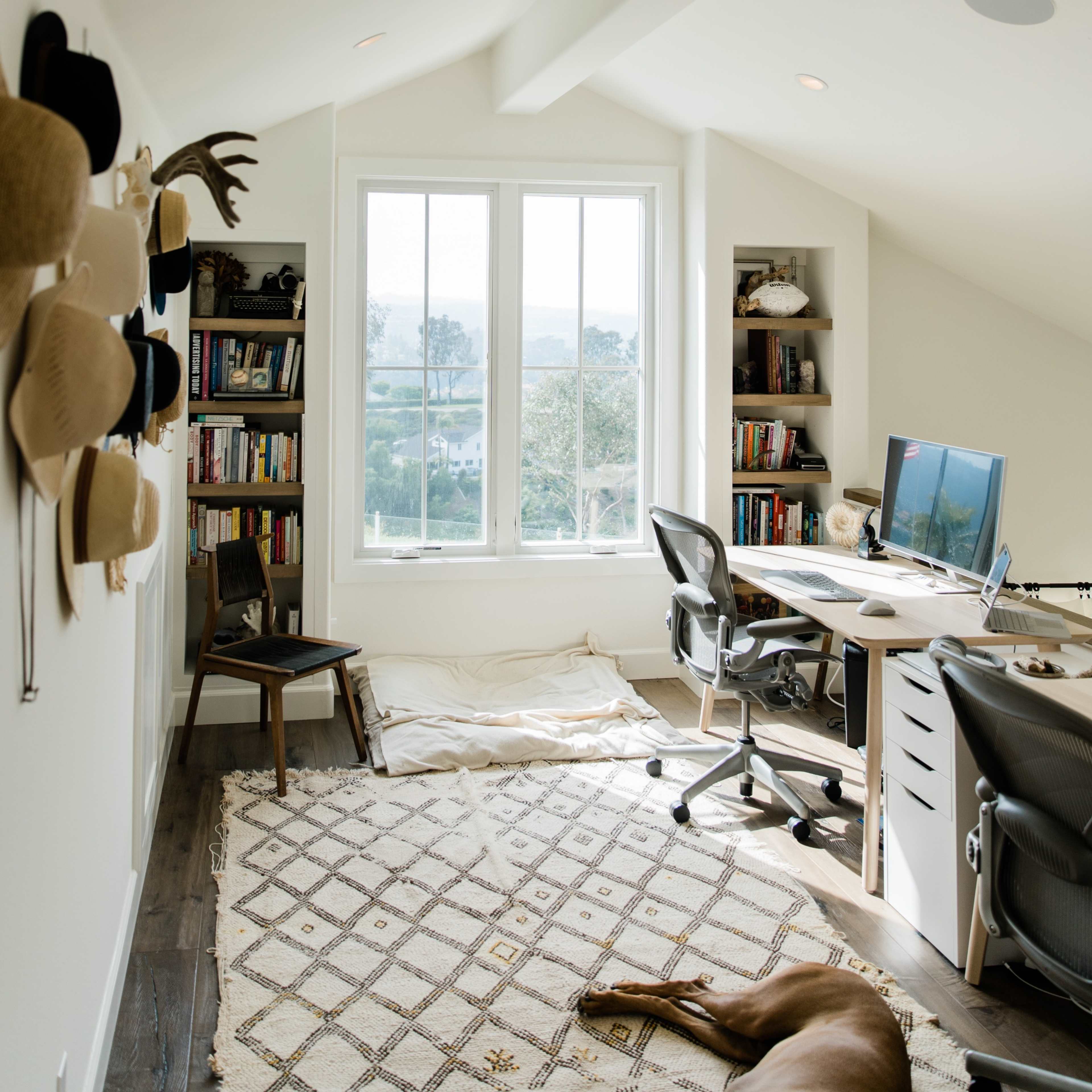 A cozy home office features a desk with a computer, bookshelves filled with books, and large windows allowing natural light to illuminate the space.