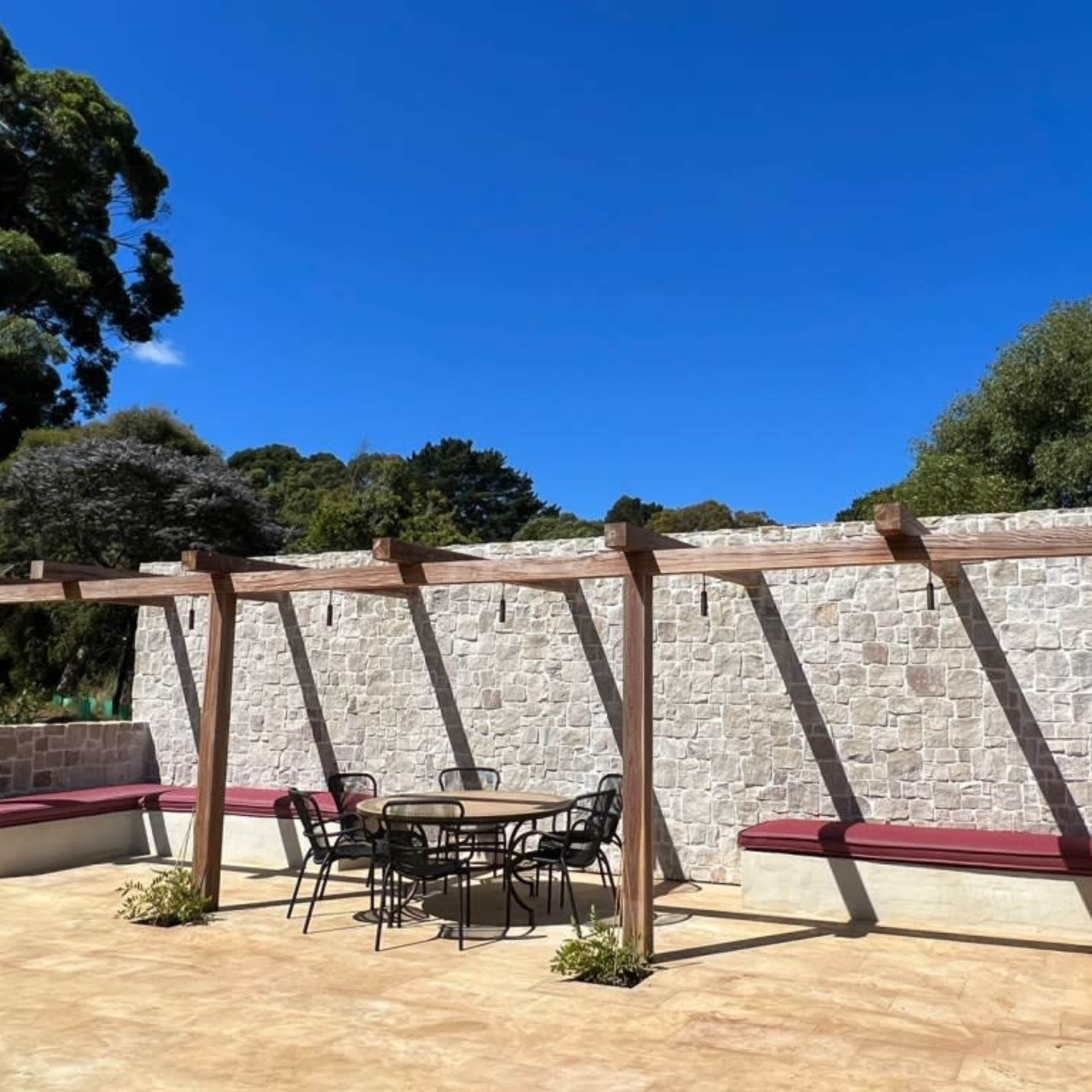 A stone wall under a clear blue sky is flanked by a wooden pergola and a table with chairs on a patio.