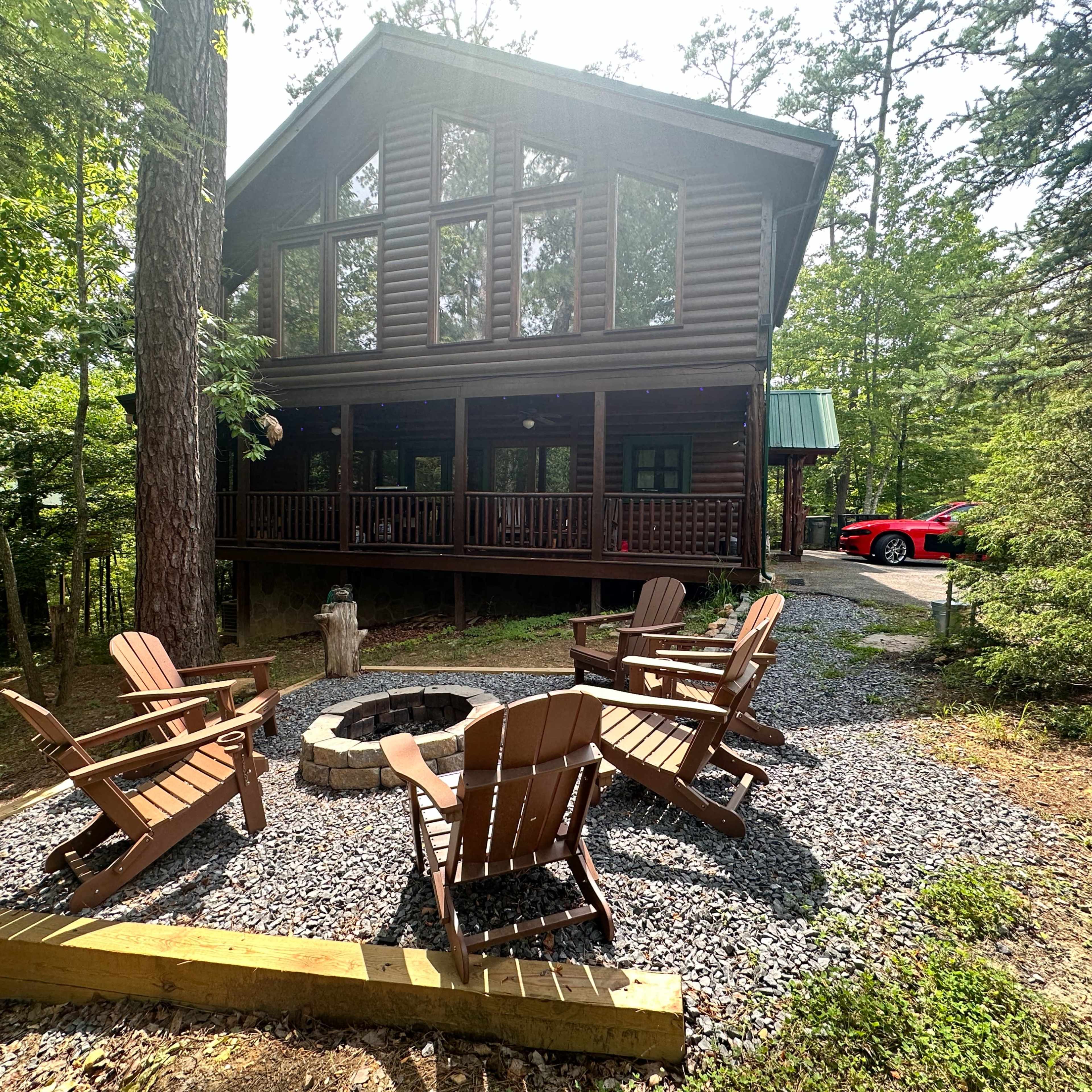 A log cabin surrounded by trees features a fire pit and several wooden chairs arranged around it, with a red car parked nearby.