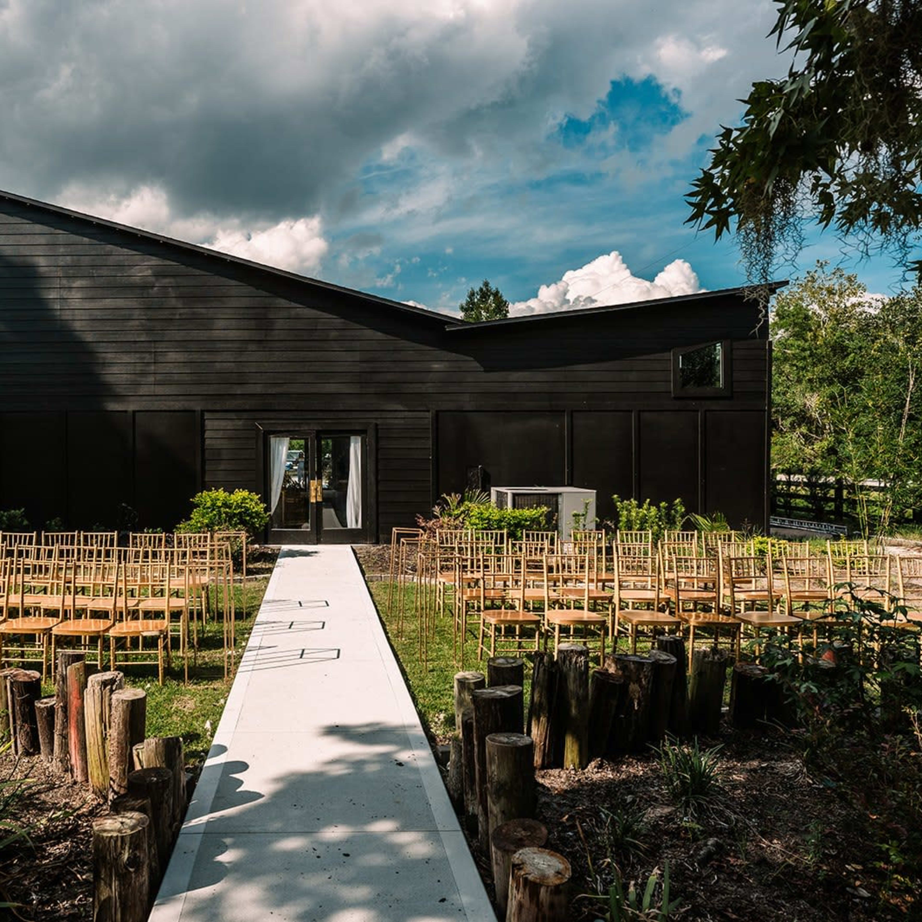 A modern black wooden building with a distinctive slanted roof, surrounded by rows of empty wooden chairs and a pathway lined with logs in a landscaped setting.