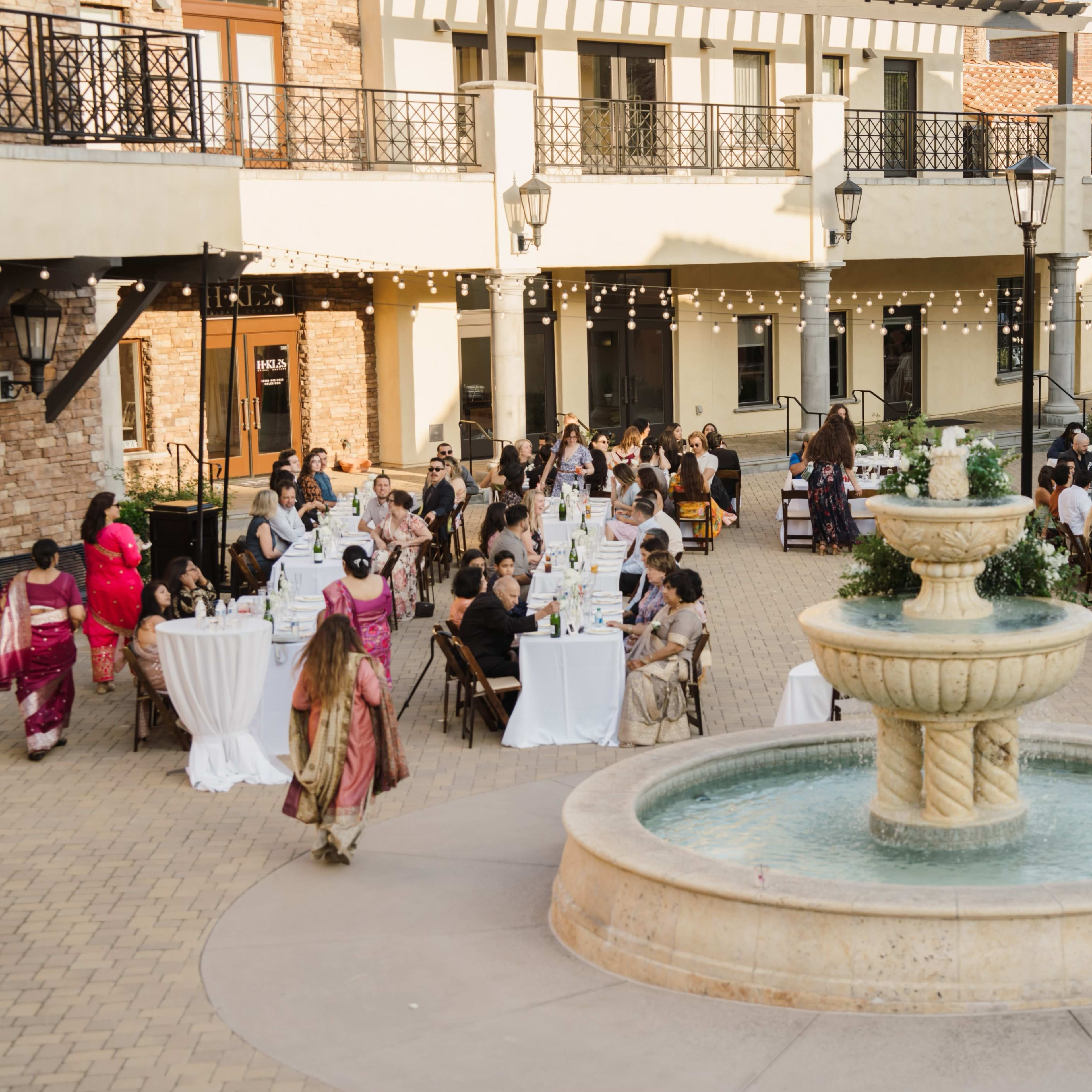 A large outdoor gathering with guests seated at tables surrounding a central fountain.