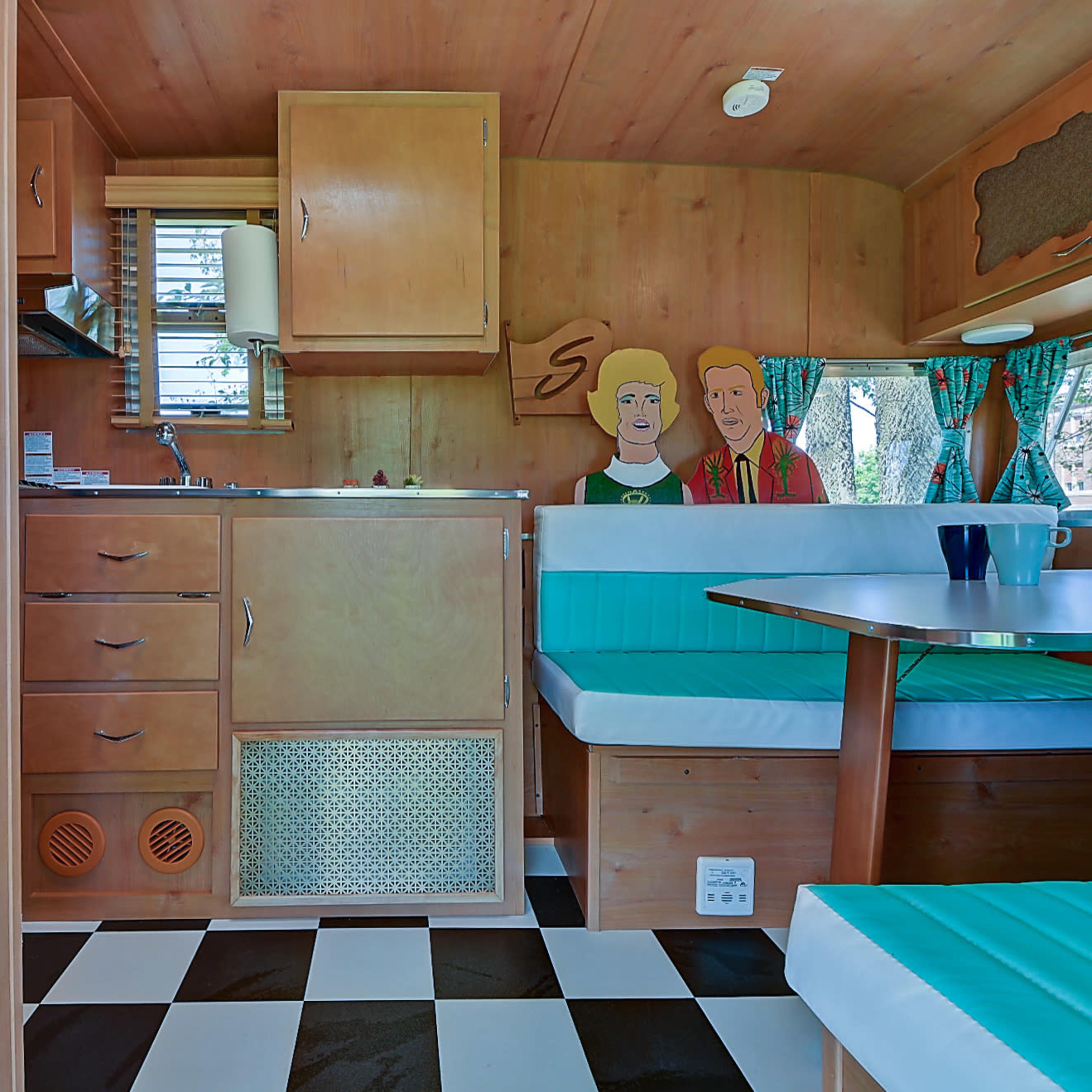 The interior of a vintage camper features wooden paneling, a kitchenette with a stove, and a dining area with turquoise seats and a checkered black-and-white floor.
