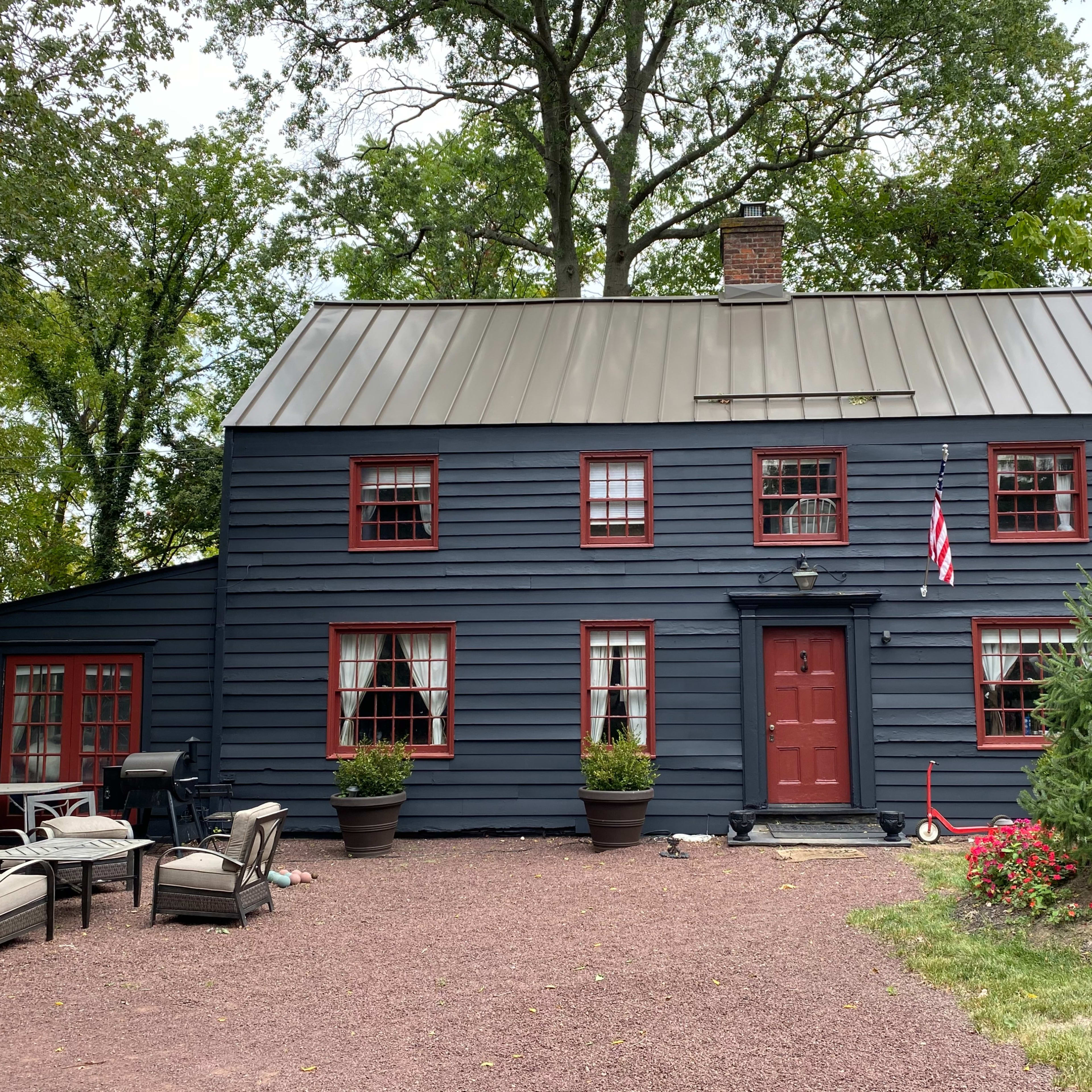 A dark blue wooden house with red trim and multiple windows, accompanied by outdoor seating arrangements and garden plants in front.