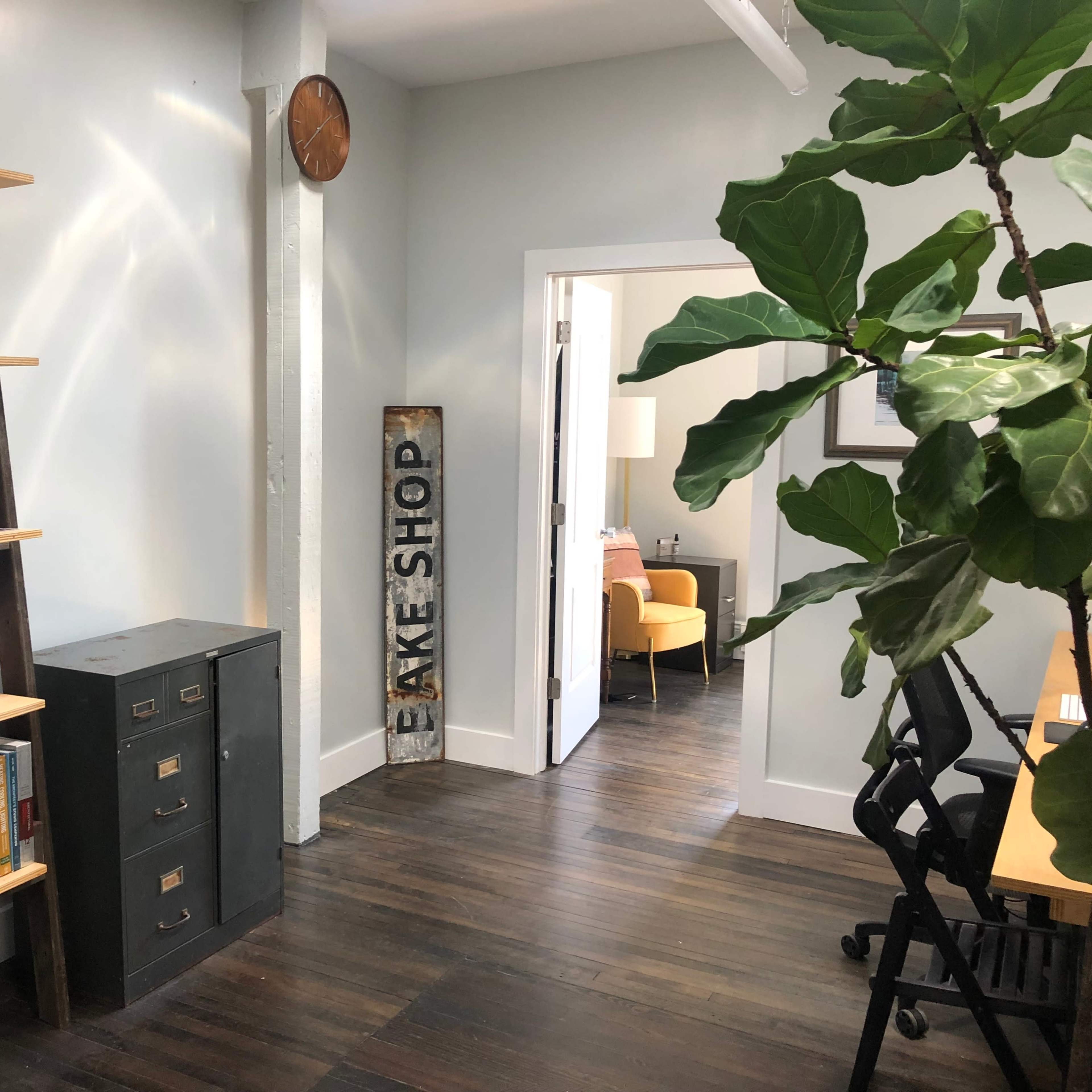 A modern interior with a wooden ladder shelf holding books, a filing cabinet, a decorative sign for a bake shop, and a doorway leading to another room furnished with a chair and lamp.