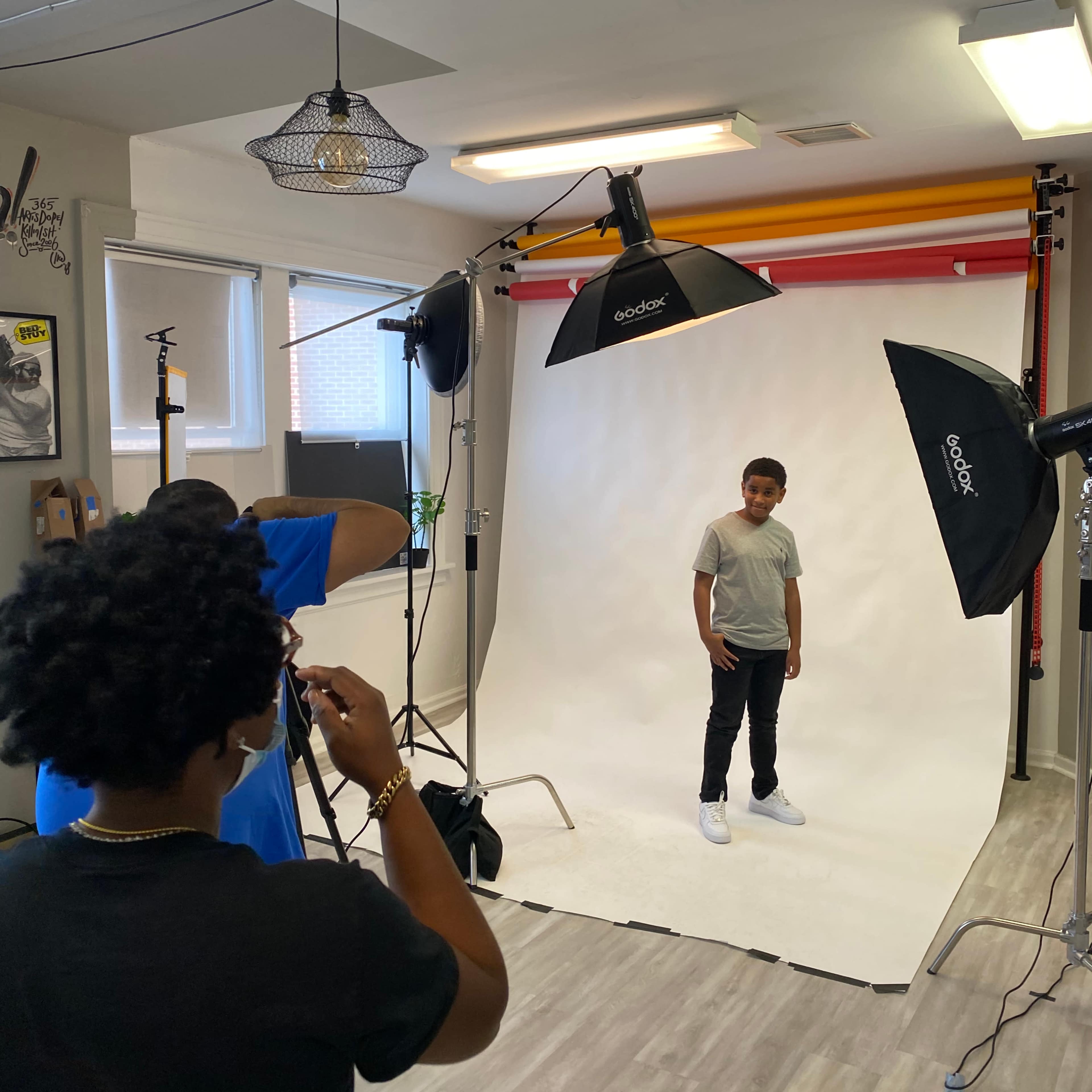 A young boy stands posing in front of a white backdrop, while a photographer adjusts equipment in a well-lit studio.