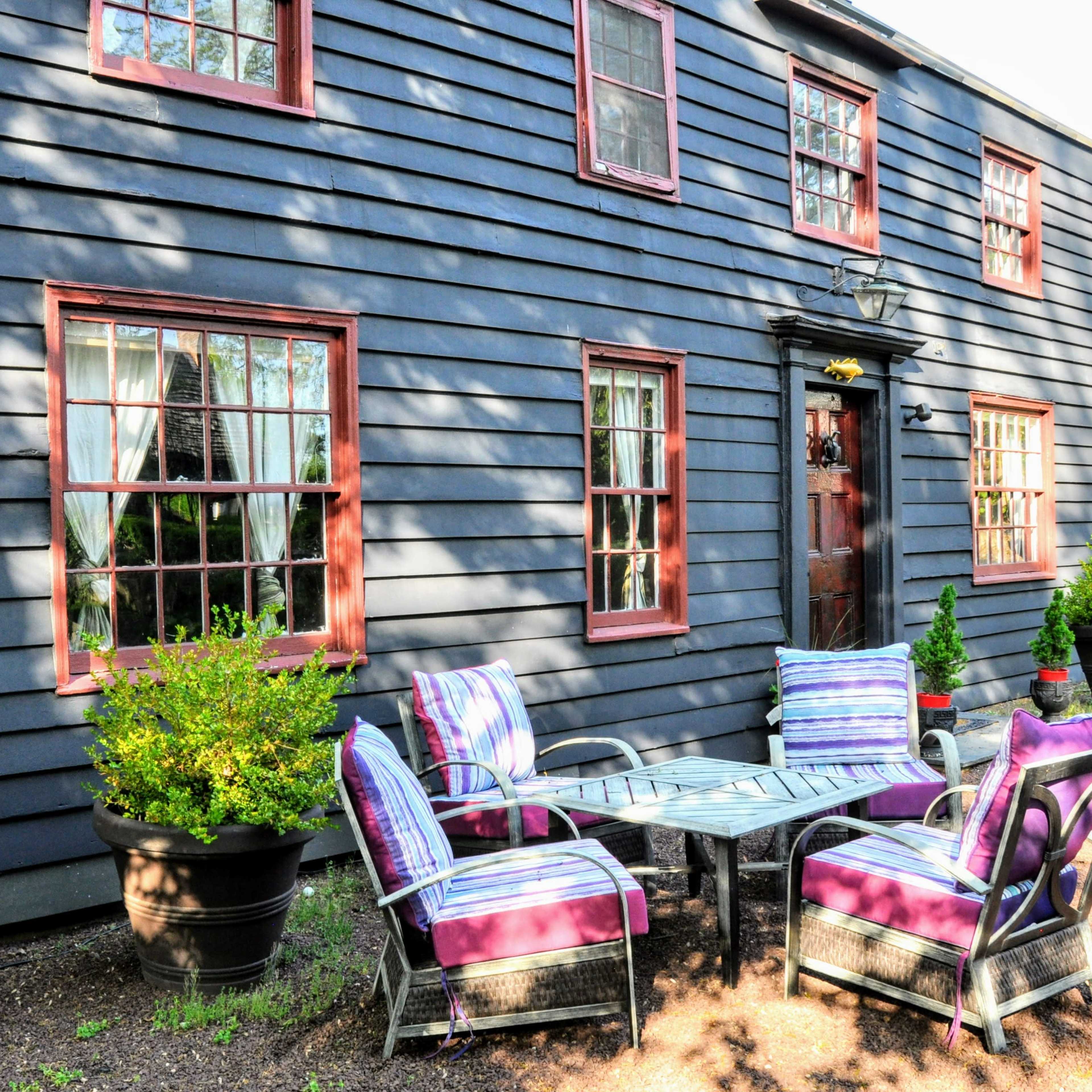 A dark wooden house with red-trimmed windows features a table and chairs on a gravel pathway surrounded by potted plants.