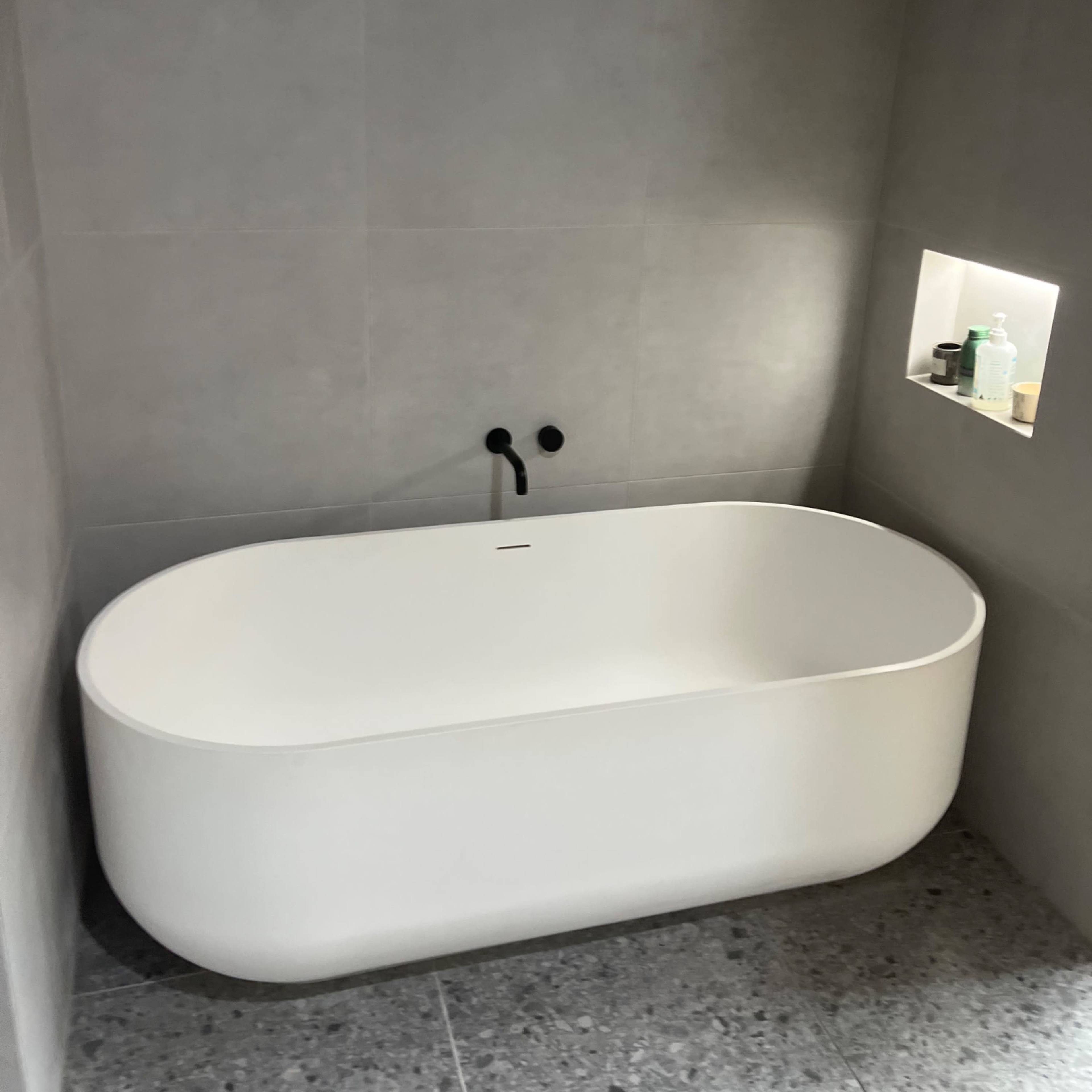 A minimalistic bathroom featuring a white freestanding bathtub against a gray tiled wall, with a small recessed nook displaying toiletries.