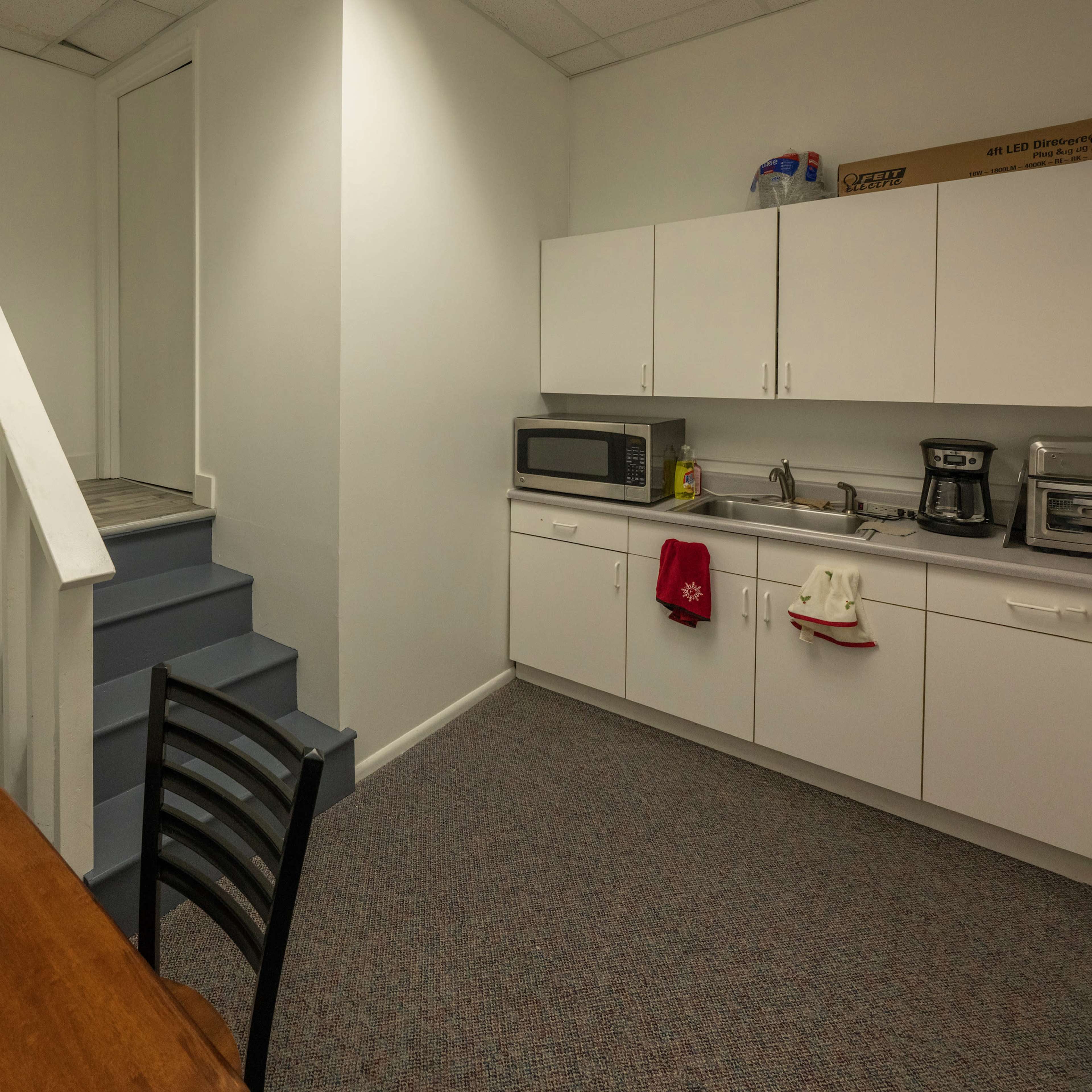 A small kitchen area with a refrigerator, microwave, and sink, next to a staircase leading to an upper level, accompanied by a wooden table and chairs.