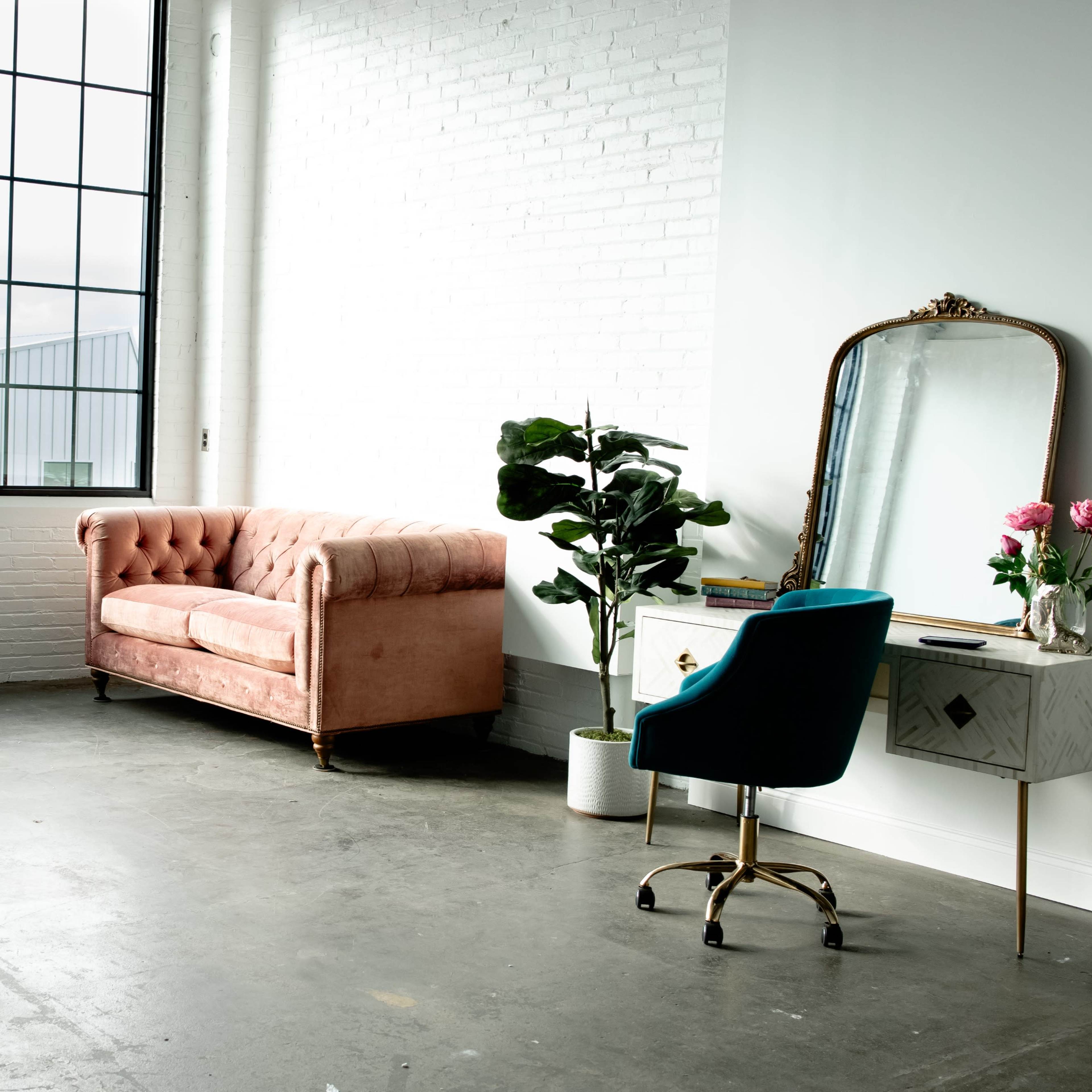 A pink tufted sofa and a small vanity desk with a chair are positioned in a bright, minimalist room with large windows and a potted plant.