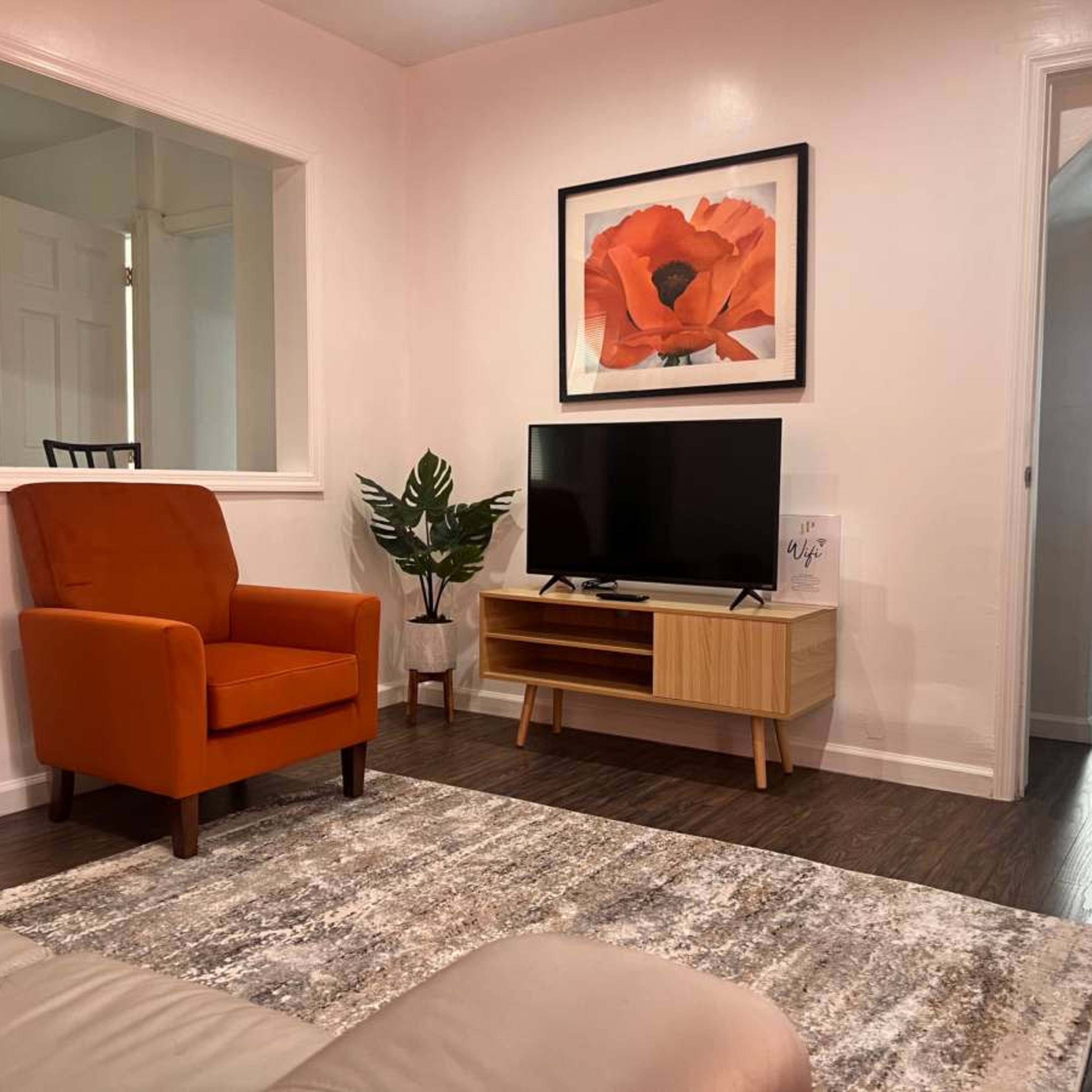 A cozy living room with an orange armchair, a television on a wooden stand, and a framed flower print on the wall.