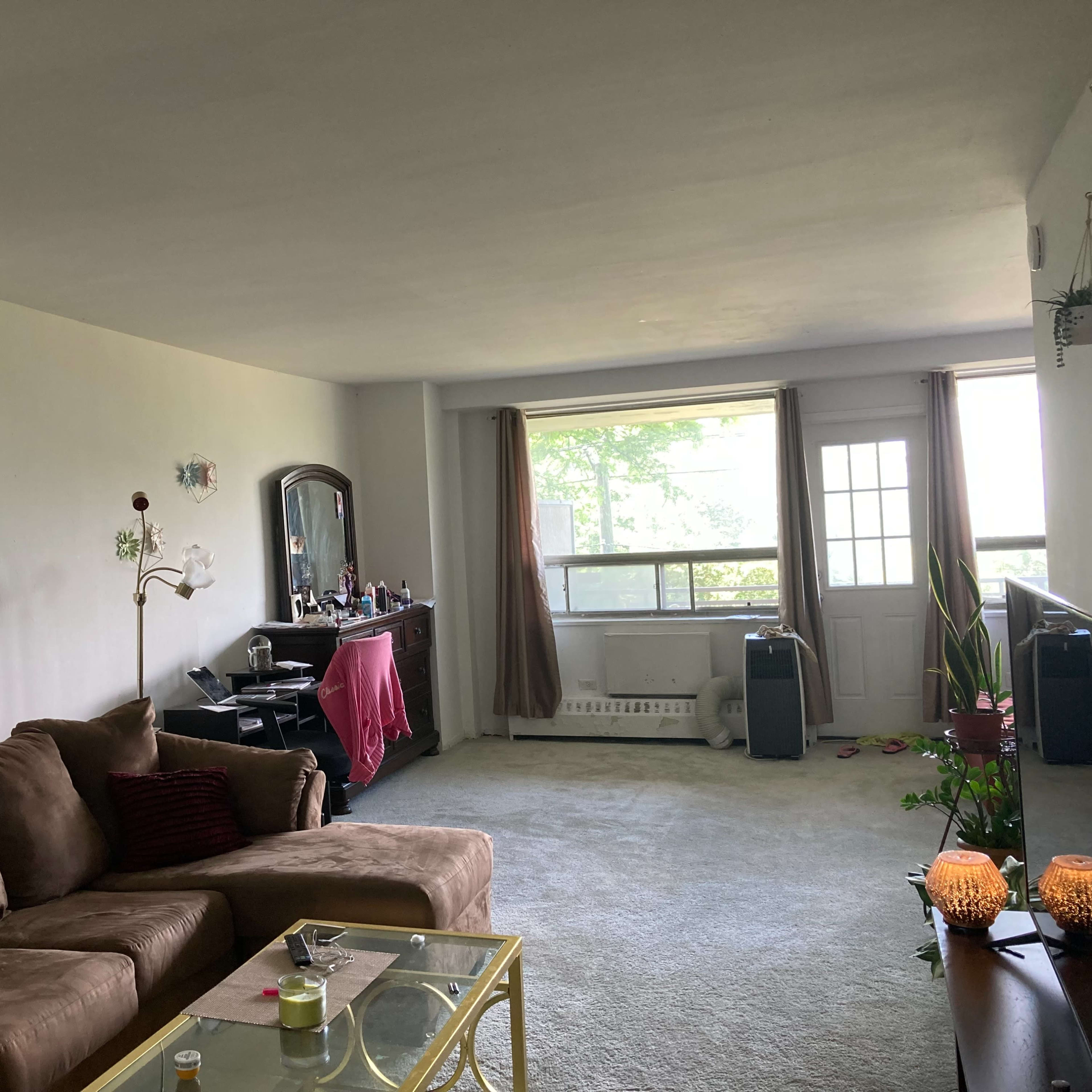 A living room with a brown couch, a glass coffee table, a mirror on the wall, and a door leading to a balcony with plants and a dresser in the background.