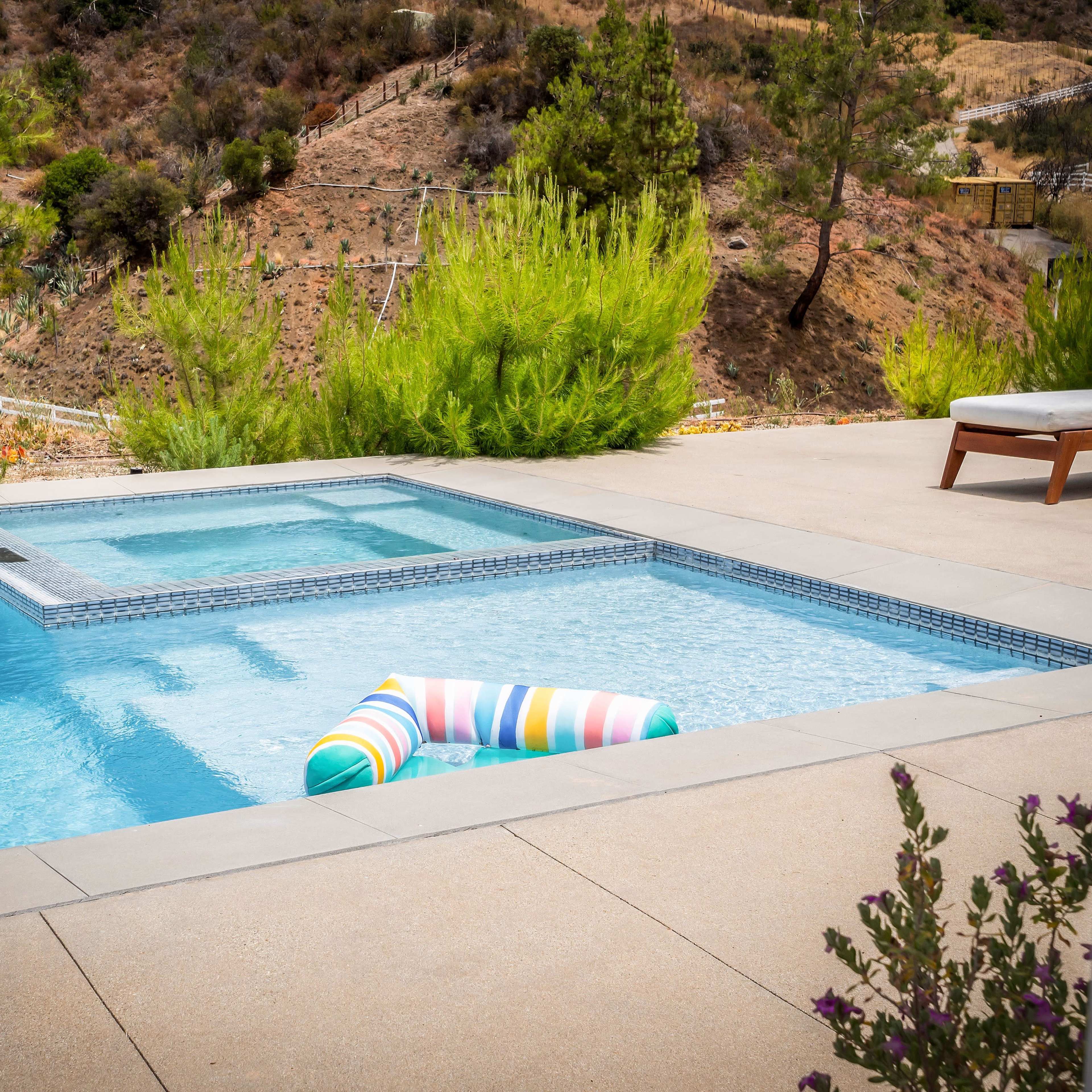 A colorful striped pool float rests in a swimming pool adjacent to a spa, surrounded by greenery and a hillside backdrop.