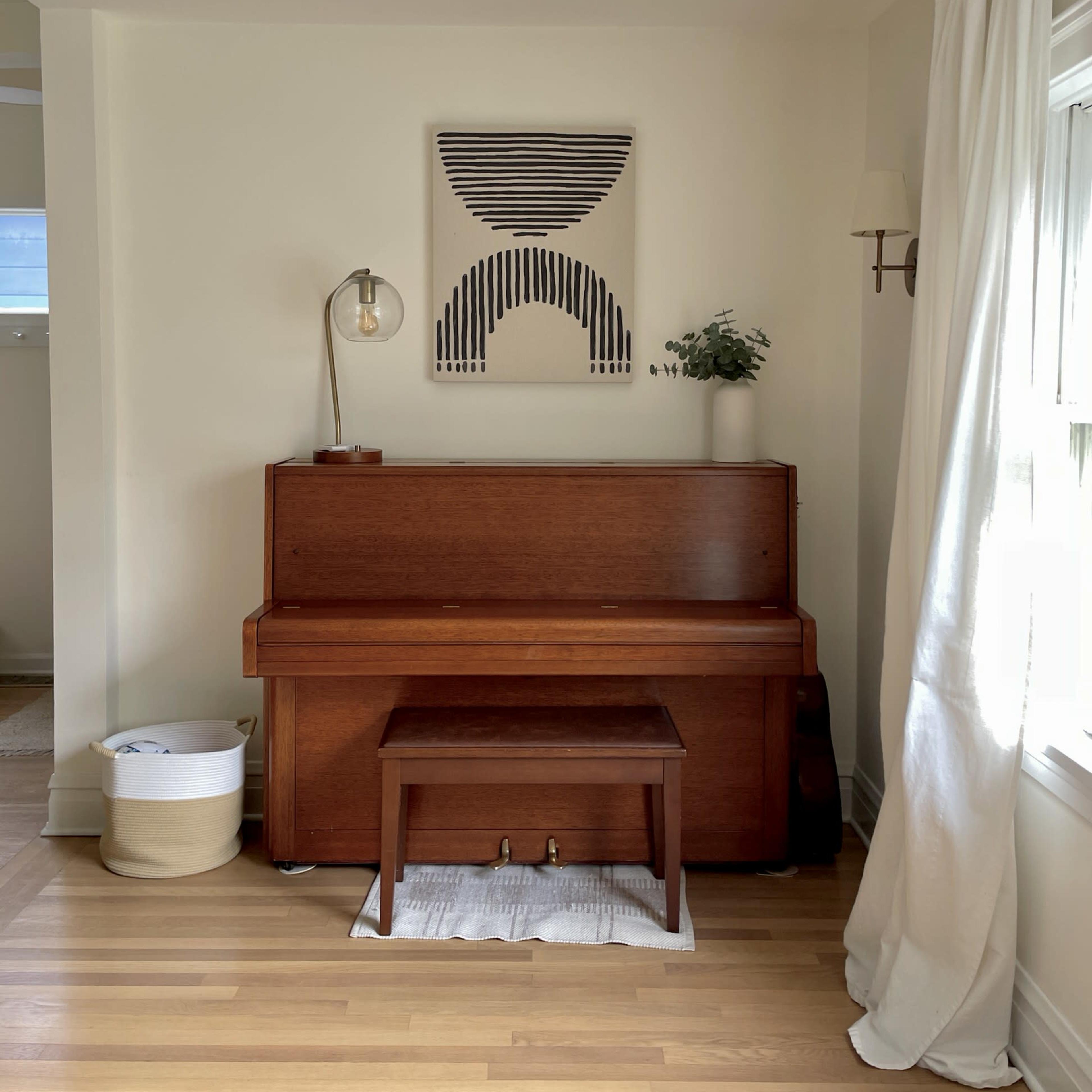 A wooden piano with a matching bench sits against a light-colored wall, decorated with a geometric artwork, and a lamp illuminates the space.