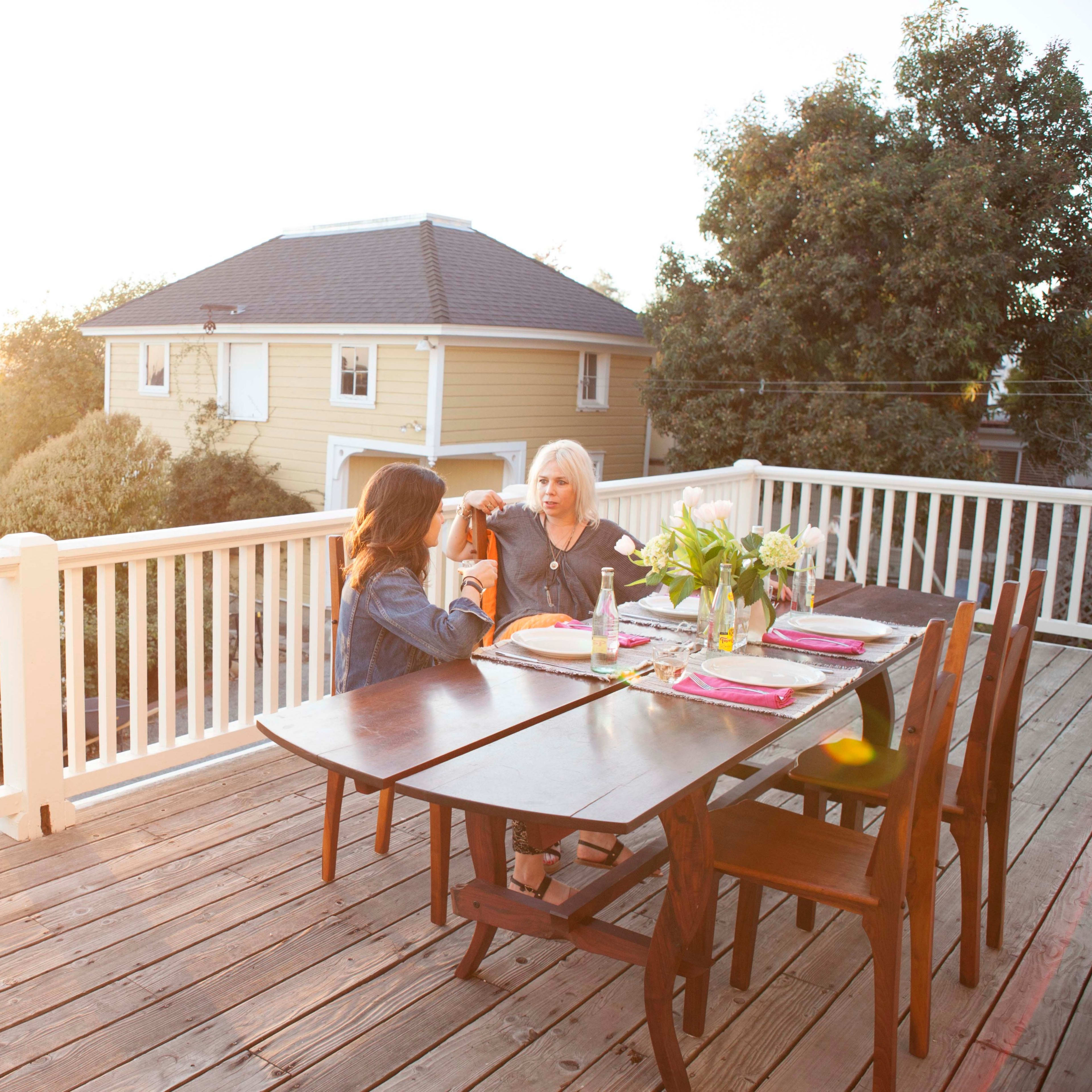 Two women sit at a wooden table on a balcony, enjoying a meal during sunset.