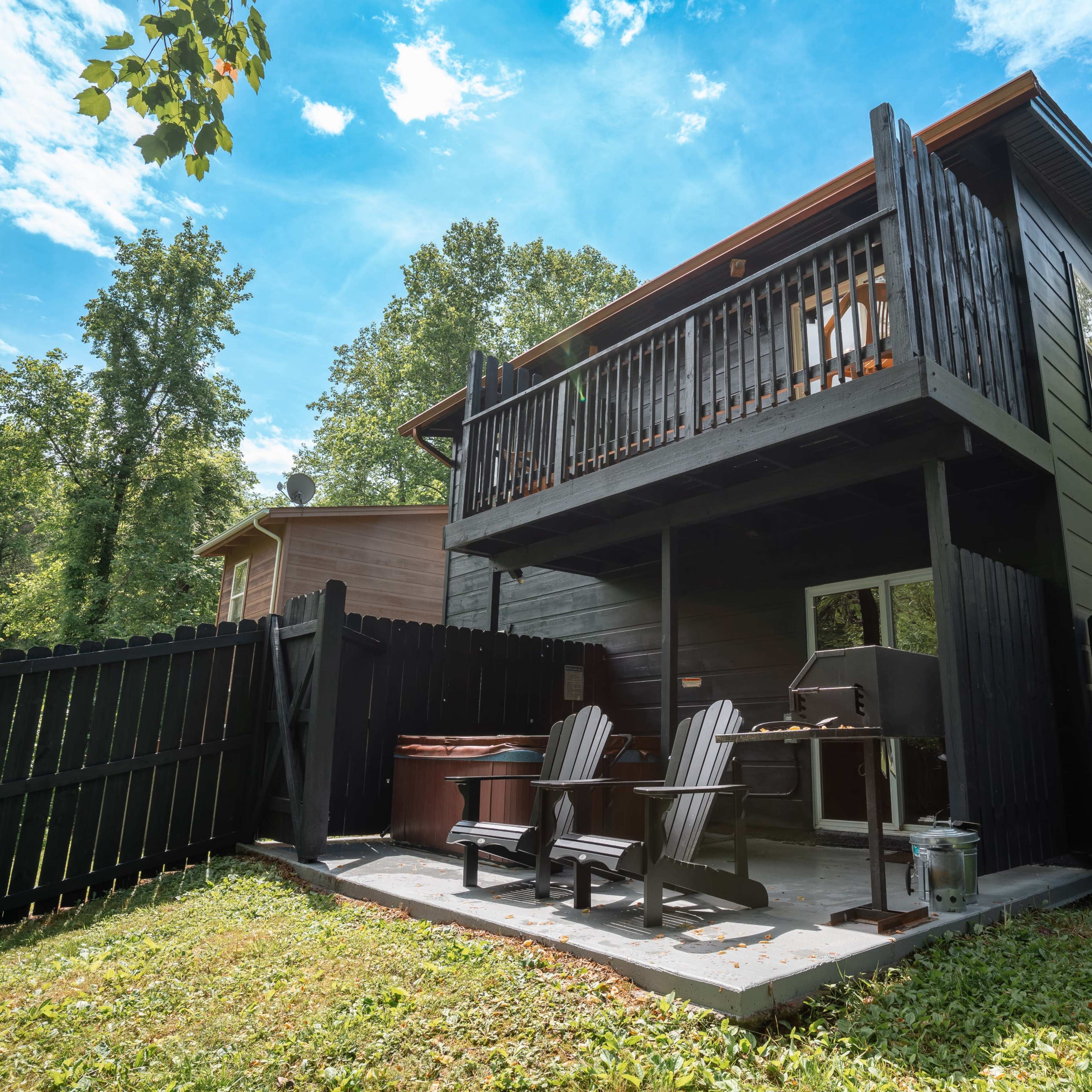 A two-story black house with a balcony overlooks a fenced outdoor area featuring seating, a grill, and a hot tub surrounded by trees.