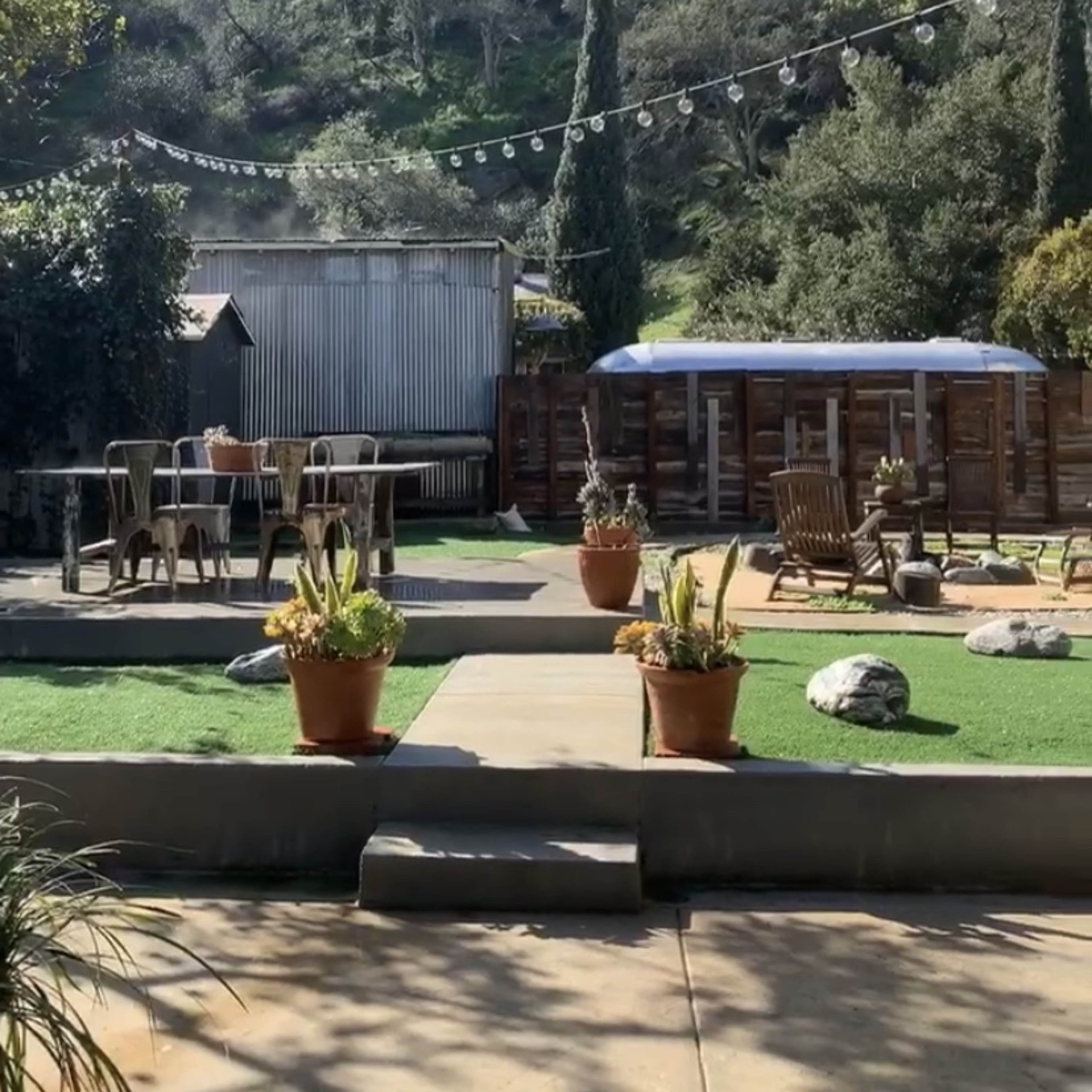 A backyard area with a patio, dining table, and planters, surrounded by greenery and hills in the background.