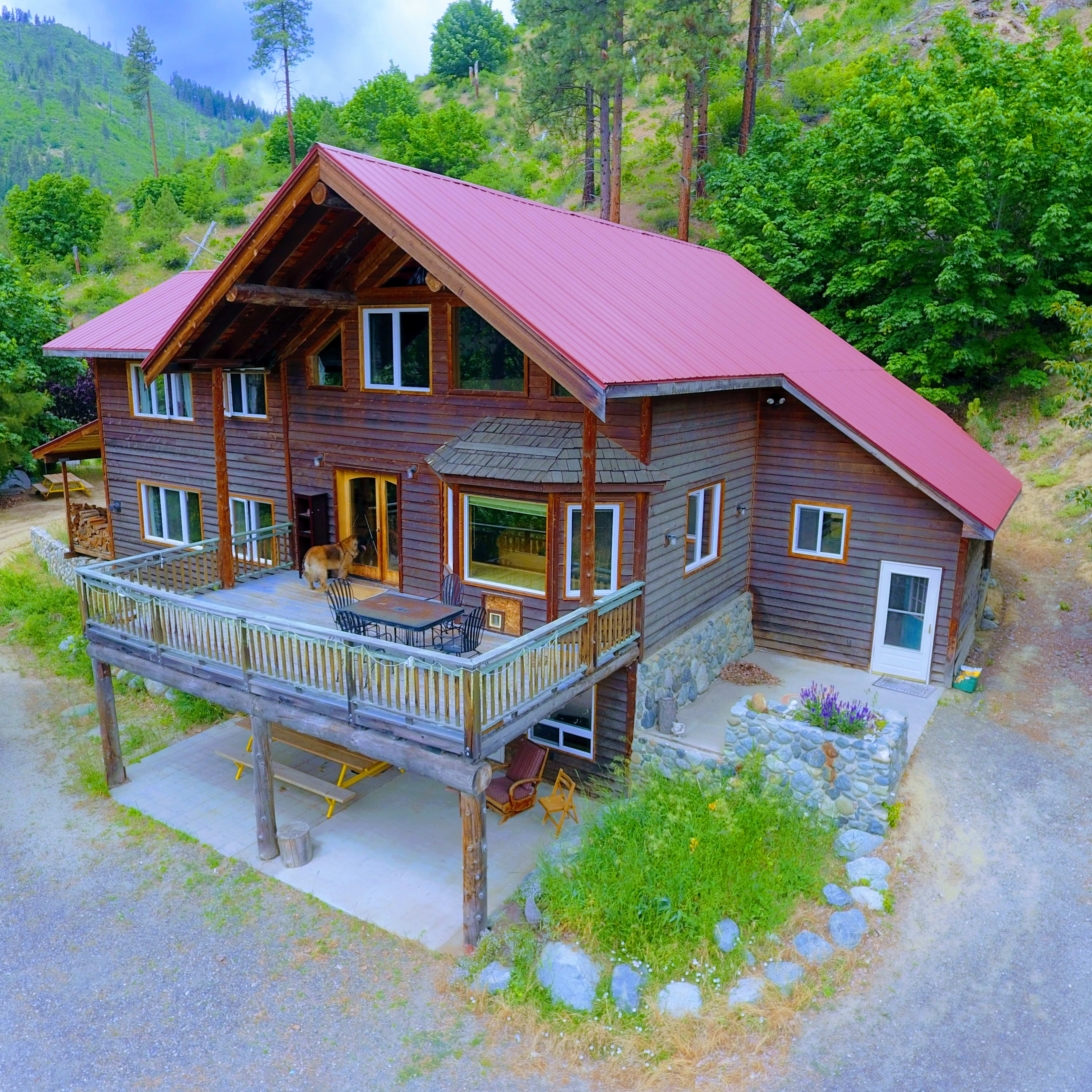 A large, two-story wooden house with a red metal roof features a spacious deck and is surrounded by trees and gravel pathways.